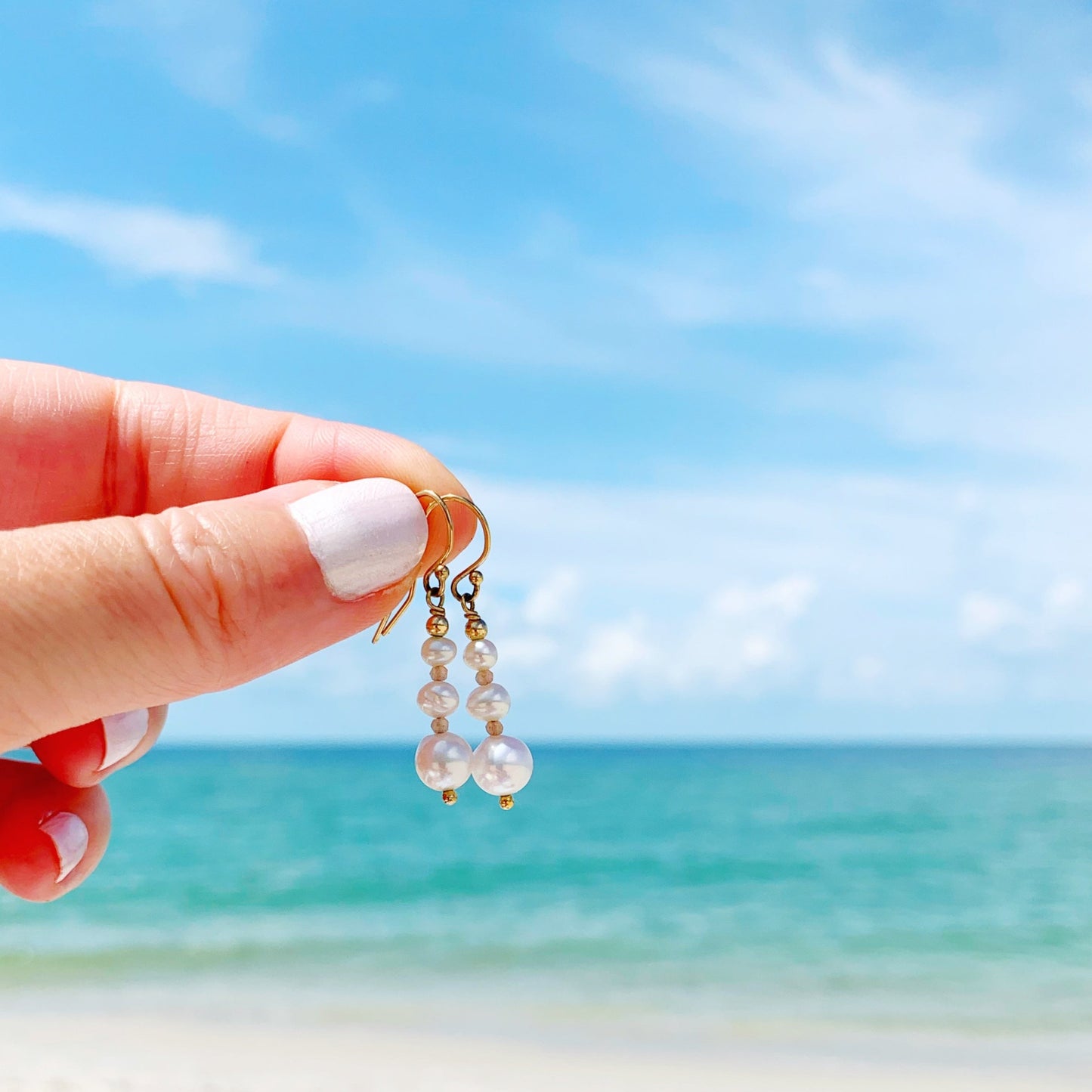 Barrington Earrings by mermaids and madeleines are created with tapered freshwater pearls and peach moonstone in linear form. this pair of earrings is held in a hand with a beach scene in the background