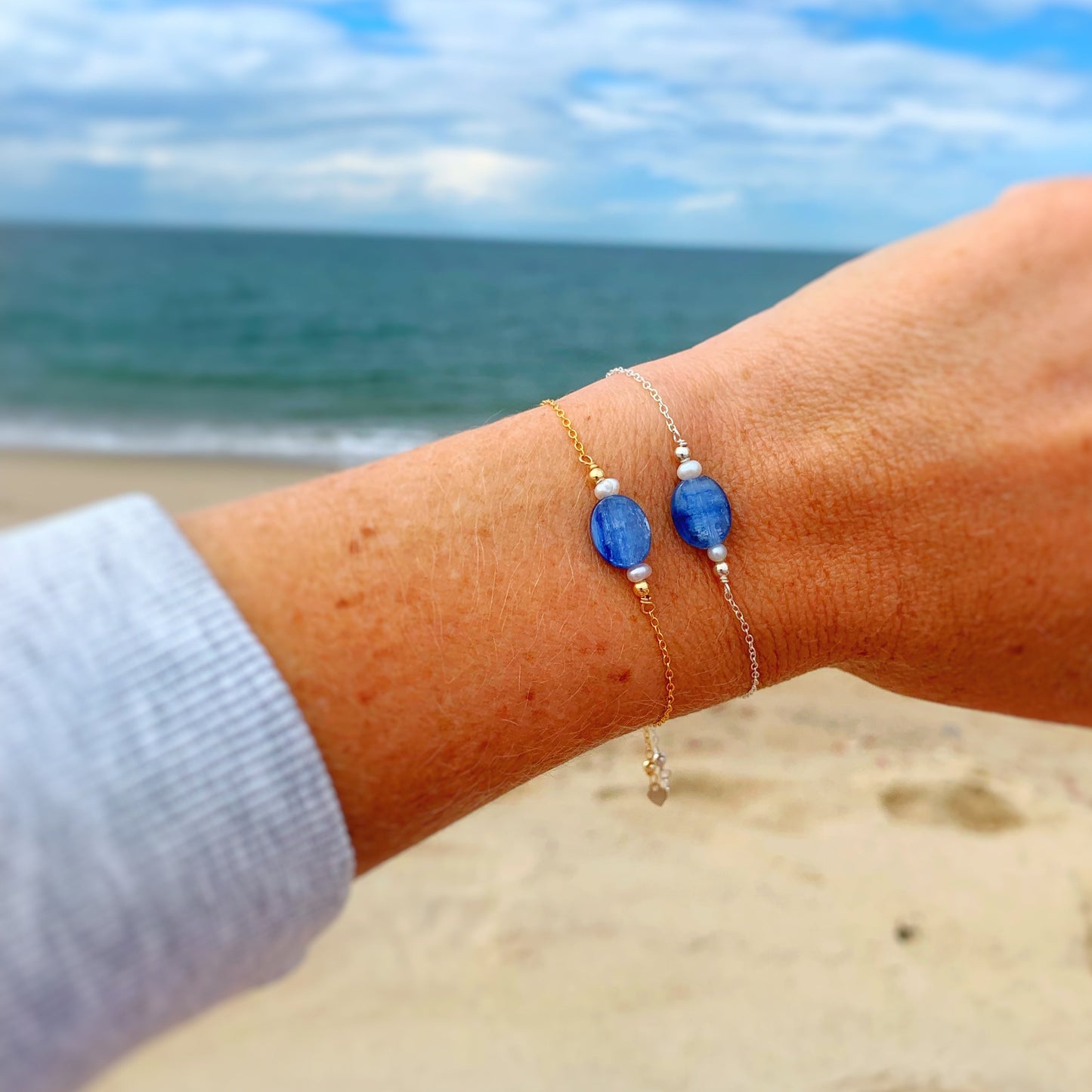 the brewster bracelet by mermaids and madeleines is an adjustable 14k gold filled chain bracelet with a slide bead at the clasp. there's a blue kyanite coin bead at the center complimented by small freshwater pearls on each side. this is a photo of the brewster bracelets in gold and silver close up on a wrist with a beach background