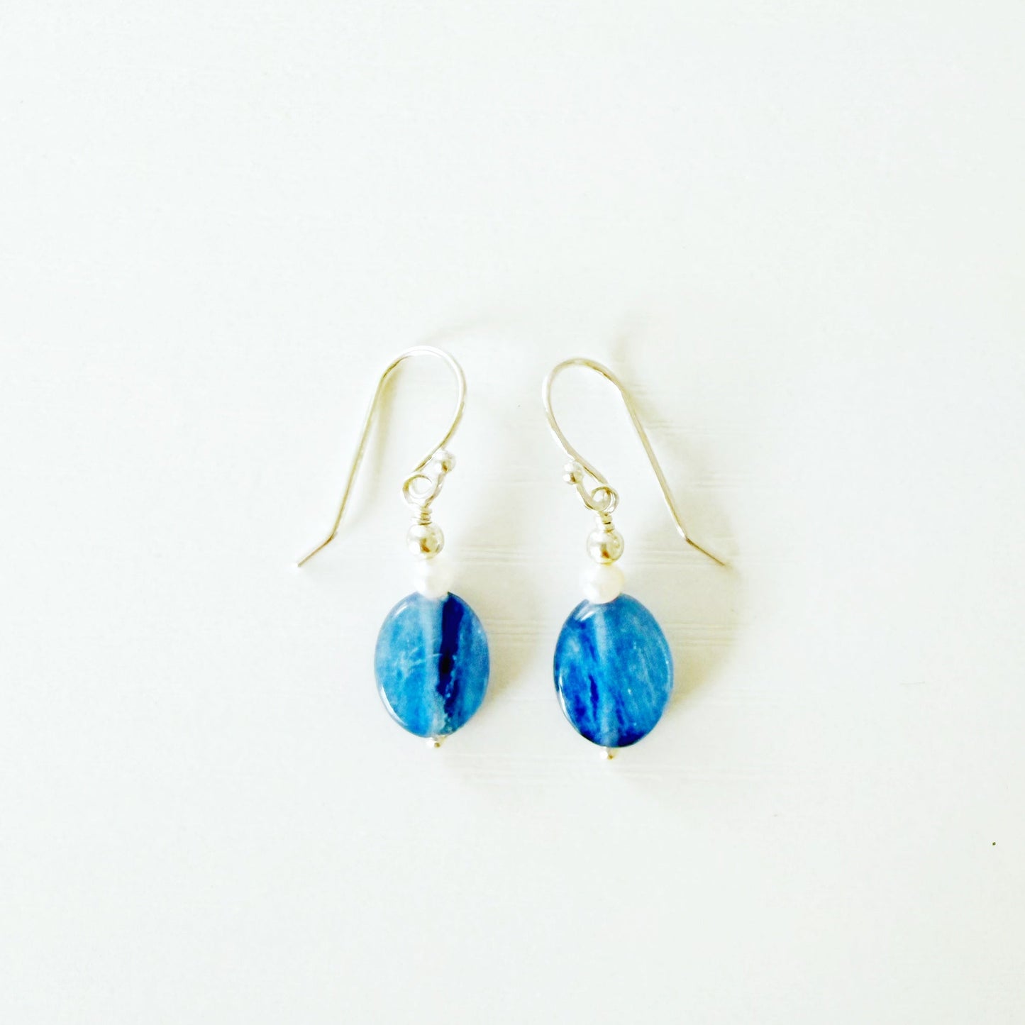 The brewster earrings by mermaids and madeleines are classic dangle style earrings with blue kyanite oval coins and tiny freshwater pearls on sterling silver earring wires. This pair is photographed flat on a white surface
