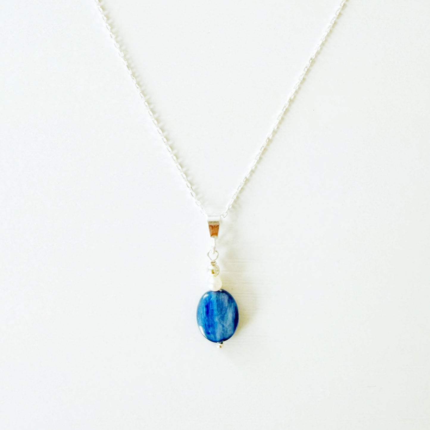 the brewster necklace by mermaids and madeleines is a classic pendant style necklace on a sterling silver chain with a blue kyanite oval coin drop and tiny freshwater pearl. this necklace is photographed close up on a white surface