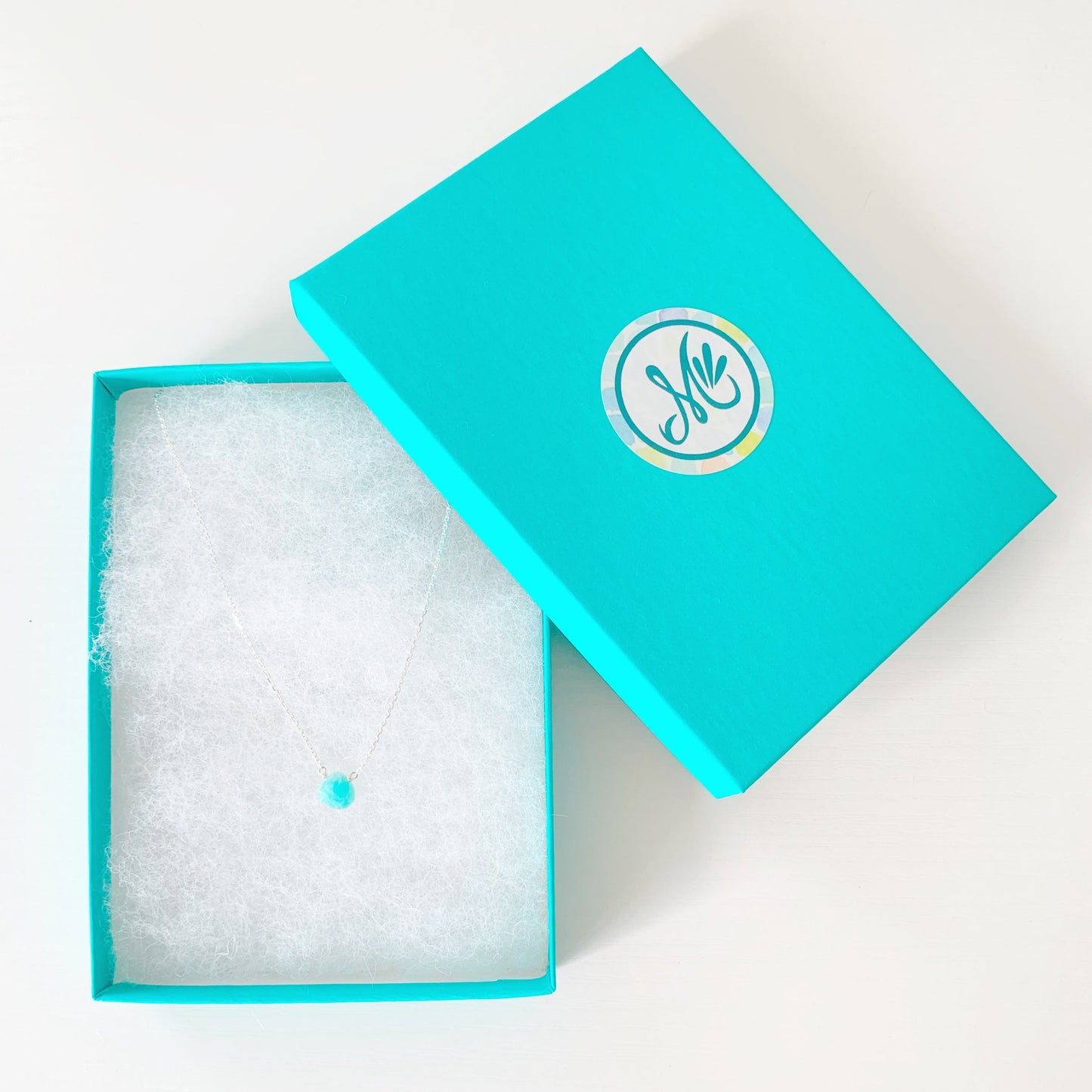 the captiva necklace by mermaids and madeleines is a dainty style necklace with a small, faceted, bright teal, amazonite briolette at the center of 17" sterling silver lightweight chain. this necklaced is placed in a teal gift box and photographed on a white surface