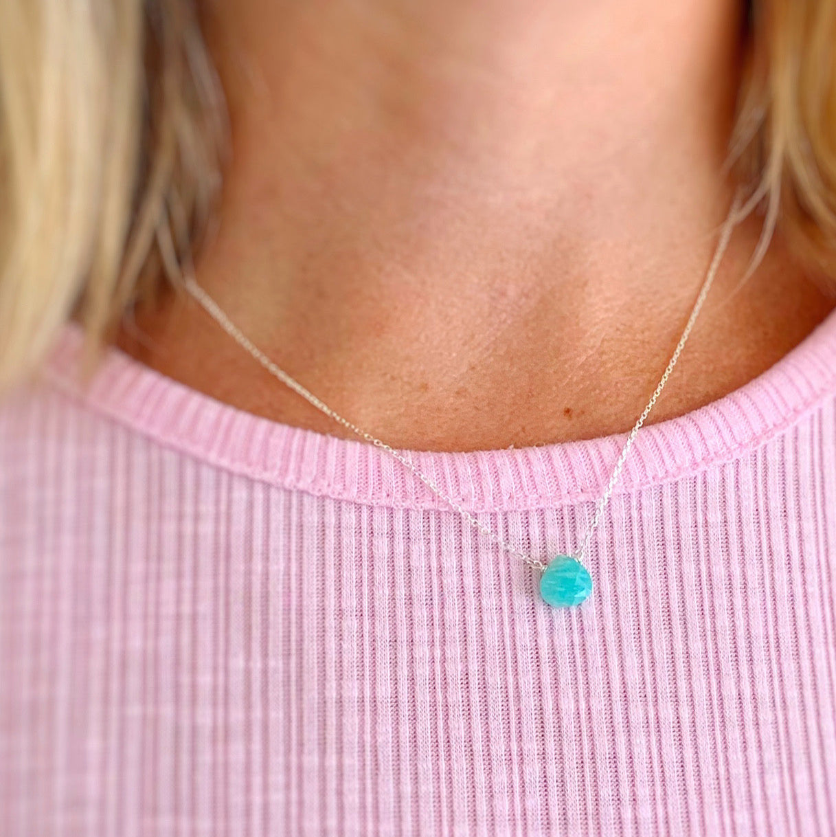The captiva necklace by mermadis and madeleines is a small faceted amazonite briolette at the center of dainty sterling silver chain. This necklace is photographed on a wearer with a light pink shirt showing the 17" length of the necklace