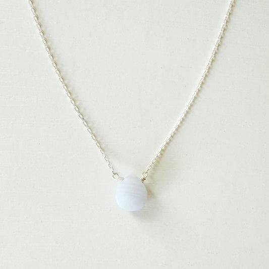 A delicate silver chain necklace with a small, smooth, pale blue-white oval pendant, displayed against a clean, off-white background.