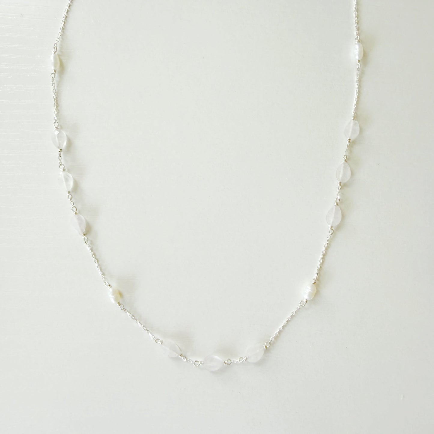 A delicate silver-toned chain necklace featuring small, irregular white pears and light pink kunzite beads spaced along the chain, displayed against a plain white background.