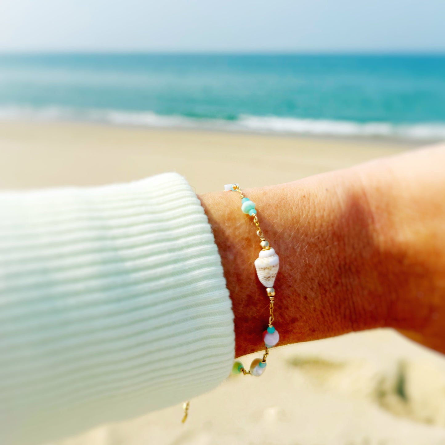 island hopper adjustable bracelet by mermaids and madeleines is a station style bracelet with a shell at the center and semiprecious beads going up the 14k gold filled chain. this bracelet is photographed close up on a wrist with a beach background