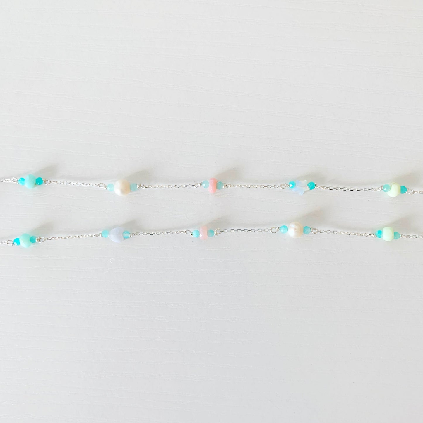 the island hopper necklace by mermaids and madeleines is a station style necklace with a shell in the center and semiprecious beads going up the sterling silver chain. this necklace is photographed showing just the semiprecious beads on the chain flat on a white surface