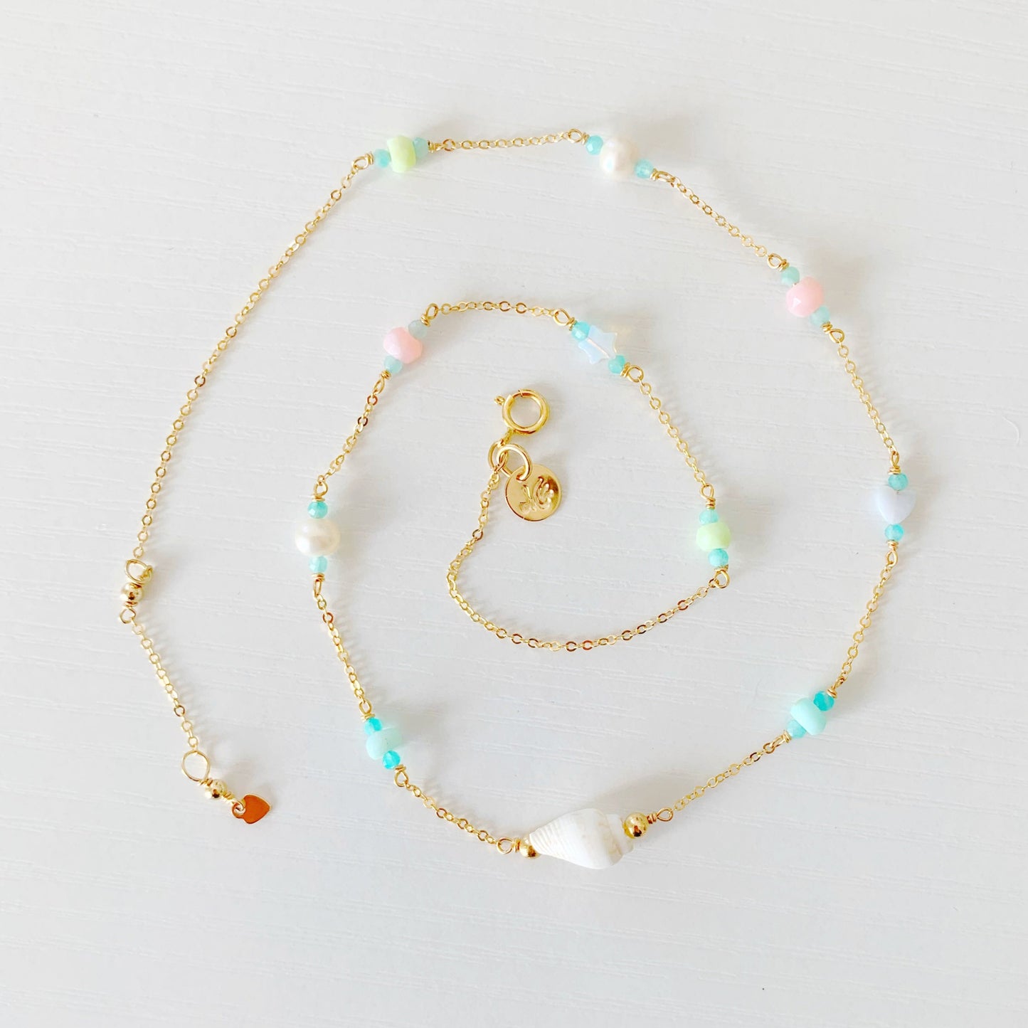 the island hopper necklace by mermaids and madeleines is a station style necklace with a shell at the center and semiprecious beads going up the 14k gold filled chain. this necklace is photographed from the top down with the necklace in a spiral shape on a white surface