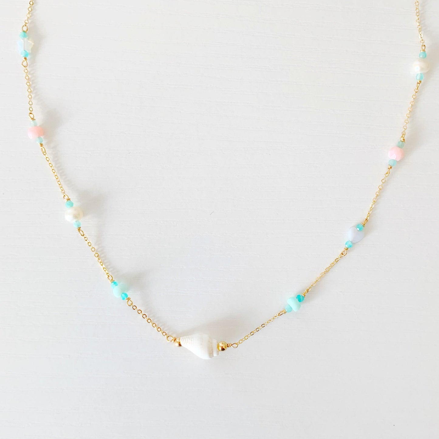 the island hopper necklace by mermaids and madeleines is a station style necklace with a shell at the center and semiprecious beads going up the 14k gold filled chain. this necklace is photographed close up on a white surface