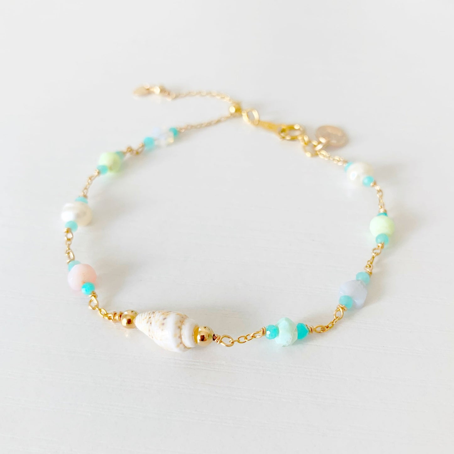 the island hopper adjustable bracelet by mermaids and madeleines is a station style bracelet with a shell at the center and semiprecious beads going up the sides of the 14k gold filled chain. this bracelet is photographed close up from the front on a white surface