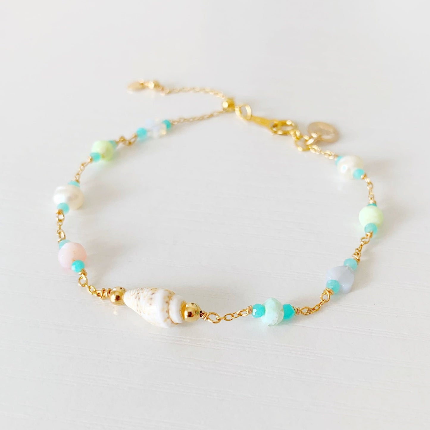 the island hopper adjustable bracelet by mermaids and madeleines is a station style bracelet with a shell at the center and semiprecious beads going up the sides of the 14k gold filled chain. this bracelet is photographed close up from the front on a white surface