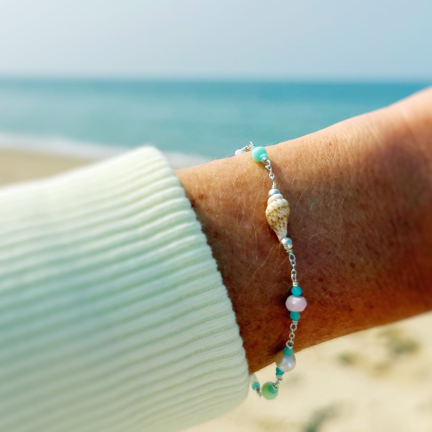 the island hopper bracelet by mermaids and madeleines is a station style necklace with a shell at the center and semiprecious beads going up the sterling chain with a slide bead at the clasp making it adjustable. this bracelet is photographed close up on a wrist with a beach background
