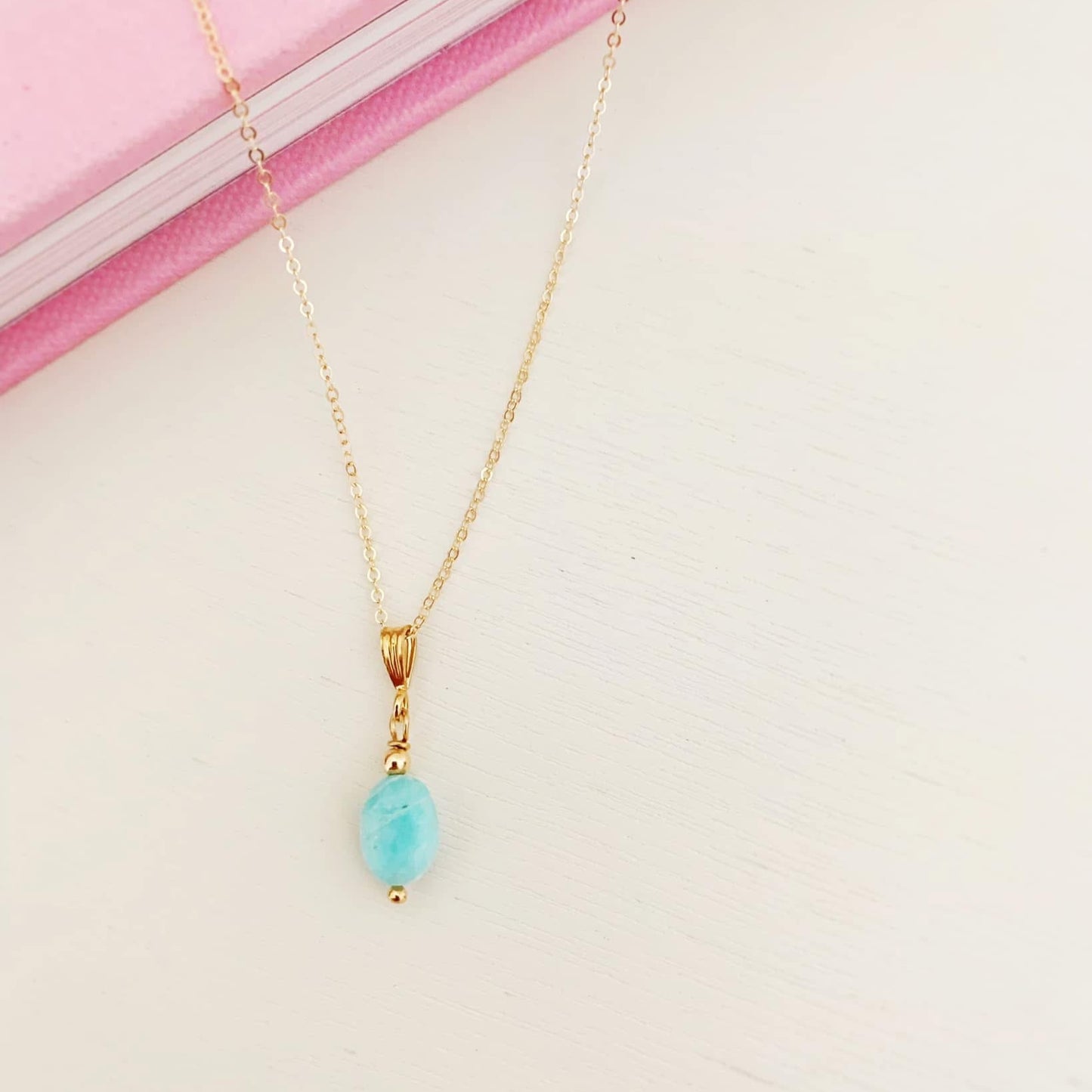 the laguna necklace by mermaids and madeleines is a pendant style necklace with a smooth, oval, amazonite drop on 14k gold filled findings and chain. this necklace is photographed on a white surface with the back half of the necklace on a pink book.