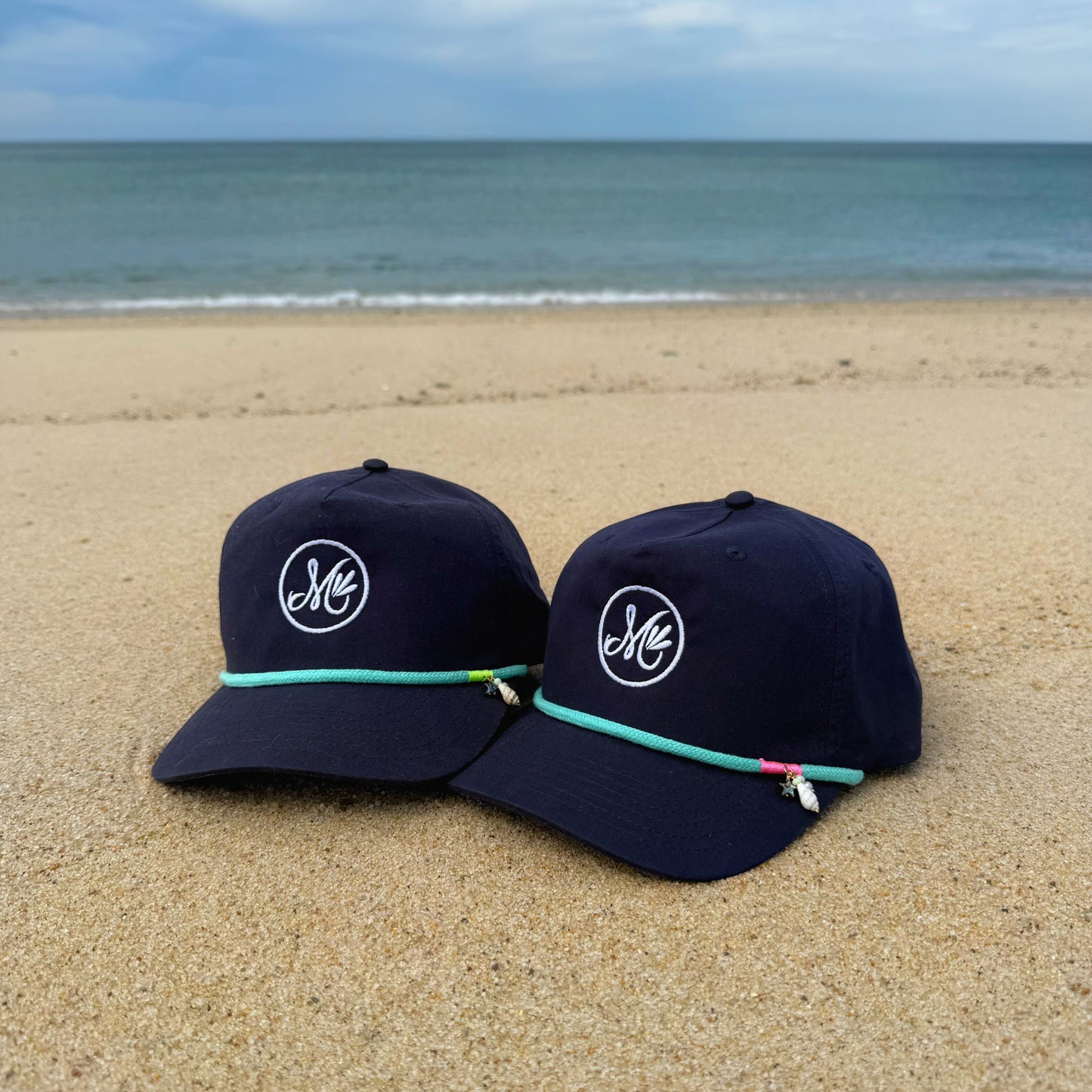 Two navy blue caps with a logo on a sandy beach with ocean in the background