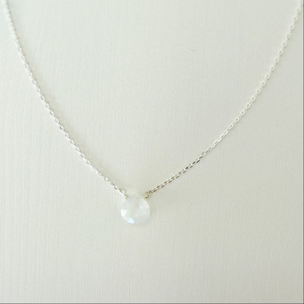a close up view of the marshmallow necklace by mermaids and madeleines which is a dainty sterling silver chain necklace with a tiny moonstone drop at the center. this necklace is photographed on a white surface