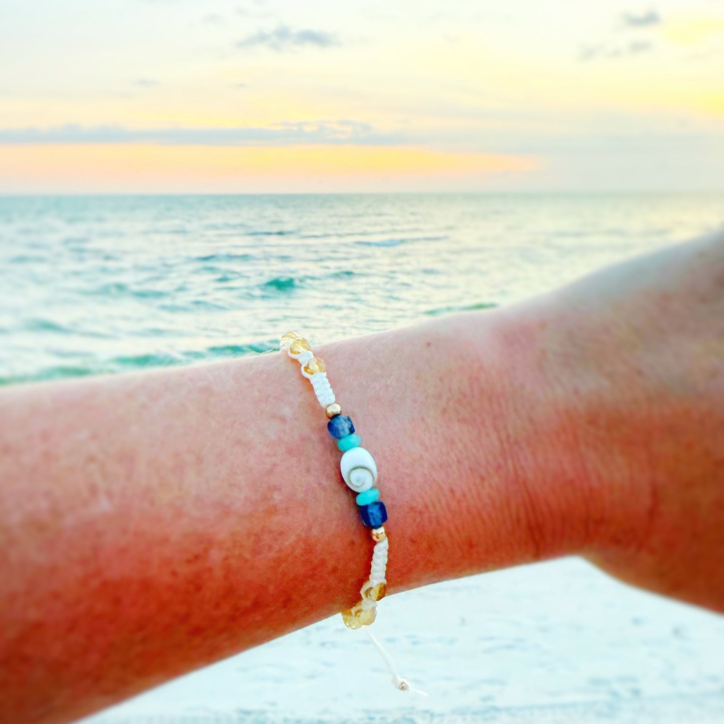 The mermaids and madeleines mermaid money macrame bracelet is created with vanilla color cord woven into a macrame frienship bracelet style. This bracelet has a shiva shell at the center and is complimented by beads of amazonite, iolite and citrine along the sides with a 14k gold filled slide bead clasp. this bracelet is photographed on an arm with a beach sunset behind it.