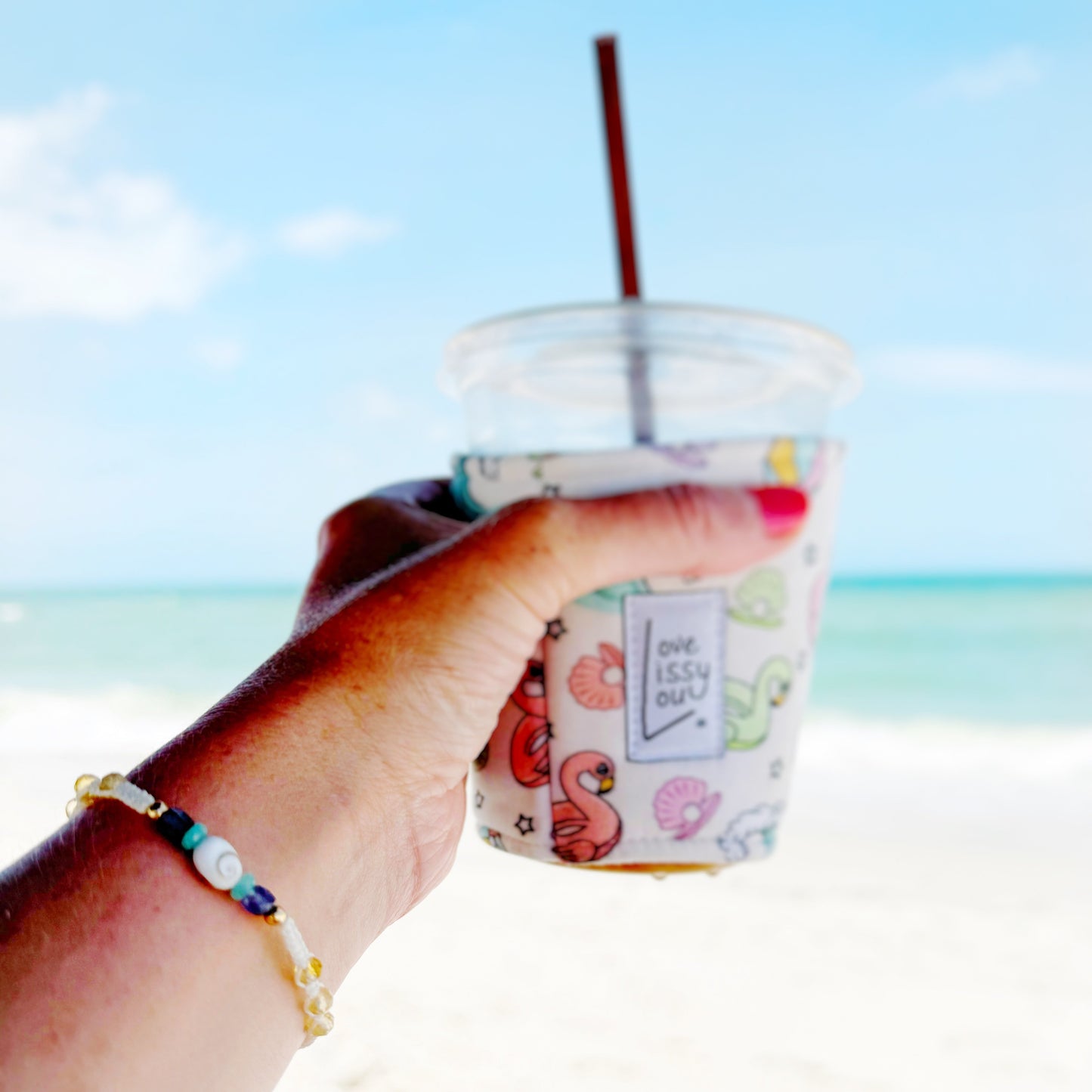 The mermaids and madeleines mermaid money macrame adjustable bracelet is created with vanilla cord with a shiva shell center and features gemstone beads of amazonite, iolite, and citrine with 14k gold filled beads and a slider clasp. This bracelet is pictured on a wrist with someone holding out an iced coffee in front of a beach scene.