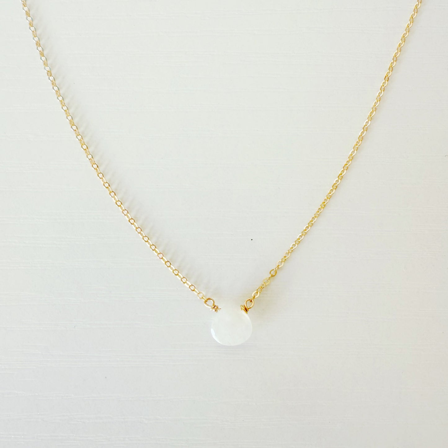the marshmallow necklace by mermaids and madeleines is a dainty 14k gold filled chain necklace with a tiny moonstone drop at the center. this necklace is photographed close up on a white surface