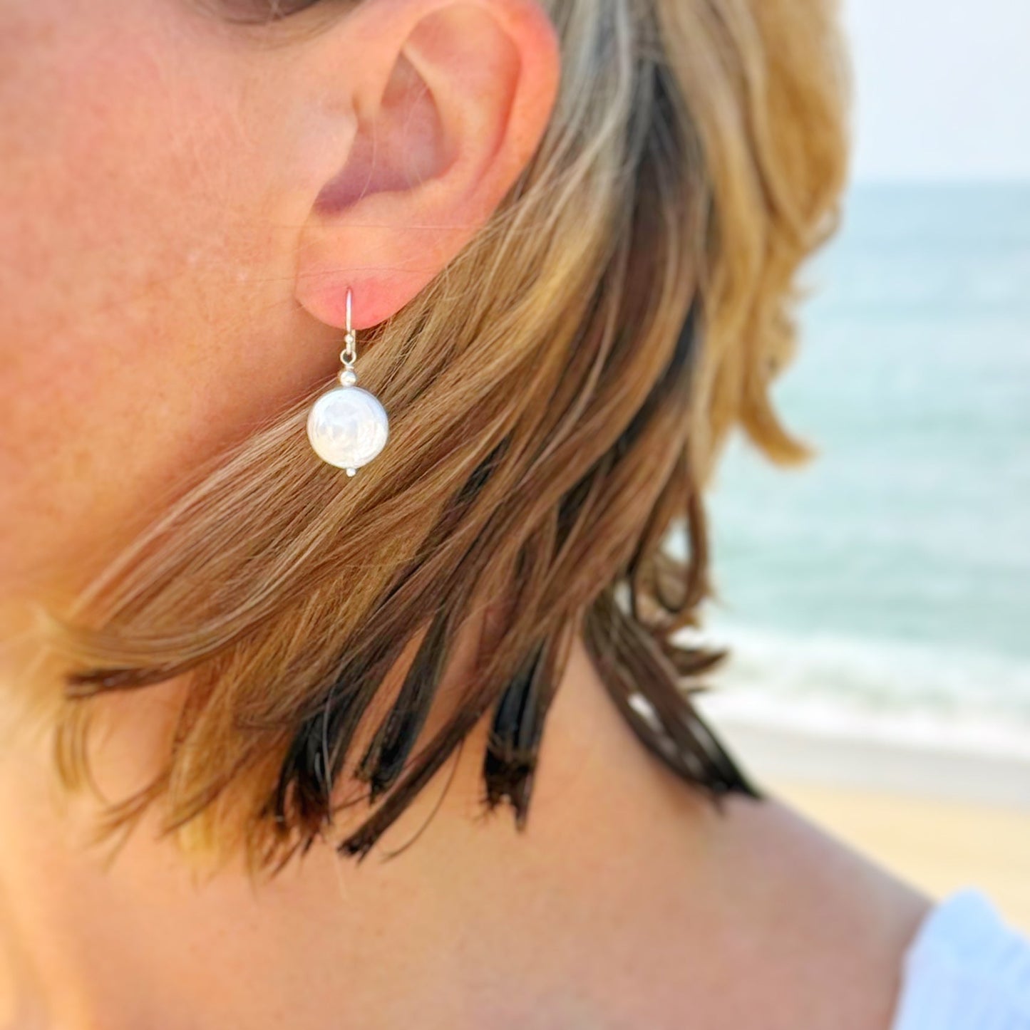 the newport earrings by mermaids and madeleines are simple pearl drop style earrings featuring a freshwater coin pearl, suspended from sterling silver ear wires and findings. A classic style that is timeless. This earring is photographed on an ear, with the wind blowing and the beach in the background