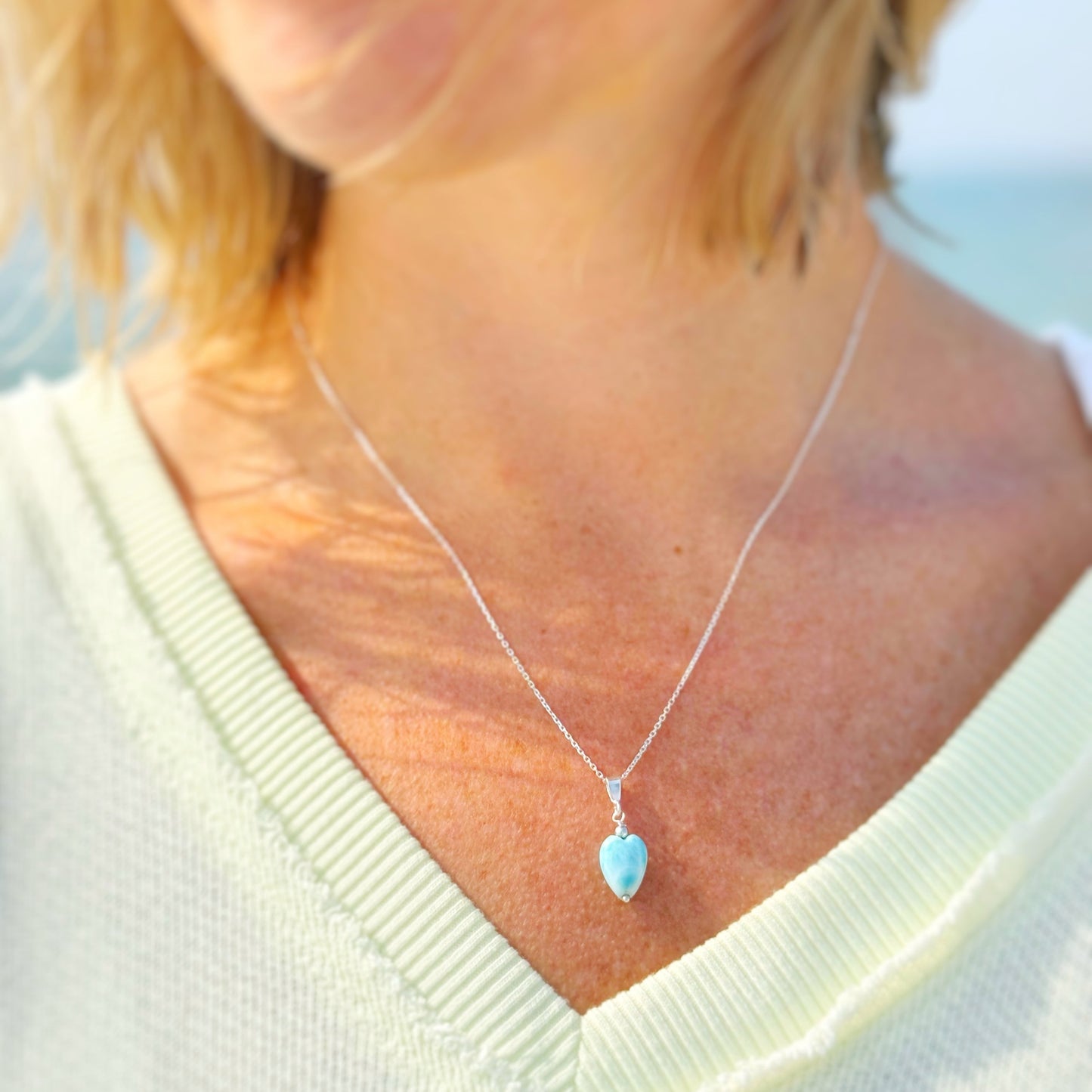 the ocean lover necklace by mermaids and madeleines is a sterling silver pendant style necklace with a heart shaped larimar bead drop. this necklace is photographed on a person close up with a beach background