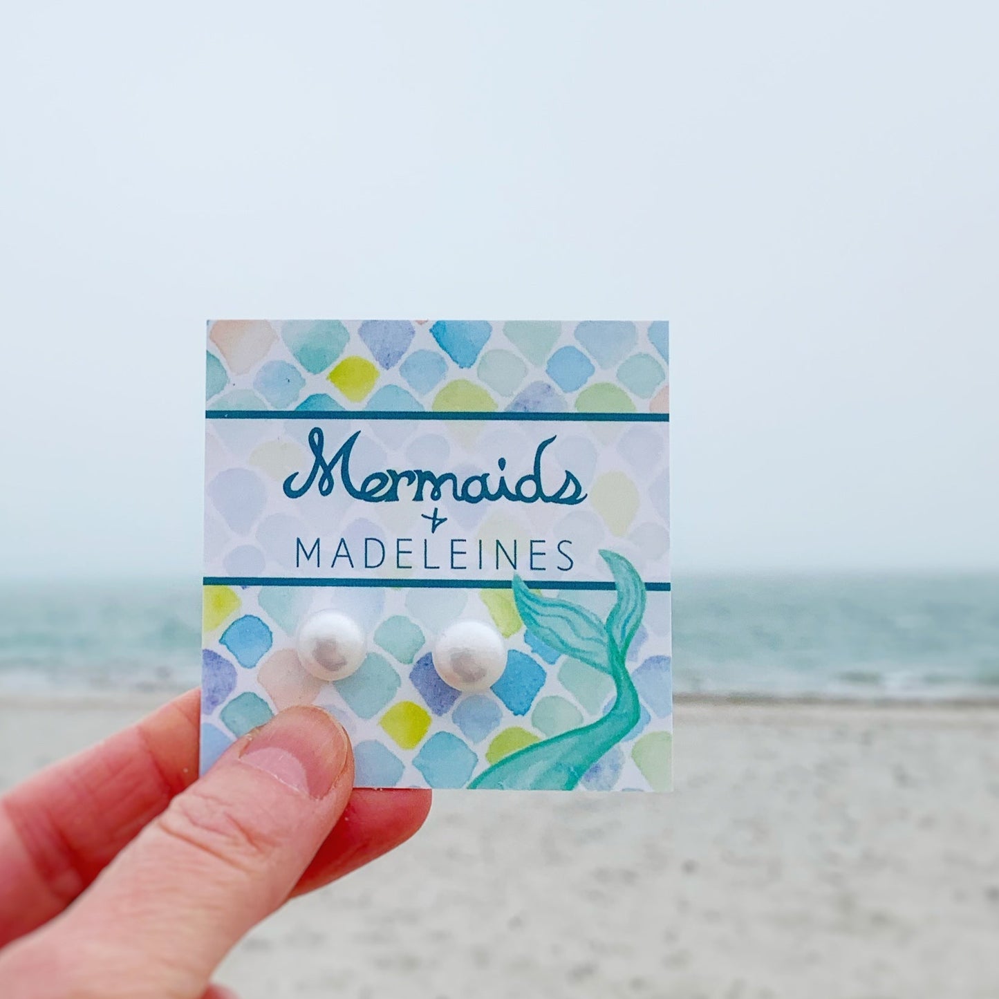 The pearlfect day pearl stud earrings by mermaids and madeleines are available in 3 sizes. this is an image of the glam size 10-11mm being held up in a hand on a branded card with a beach scene in the background
