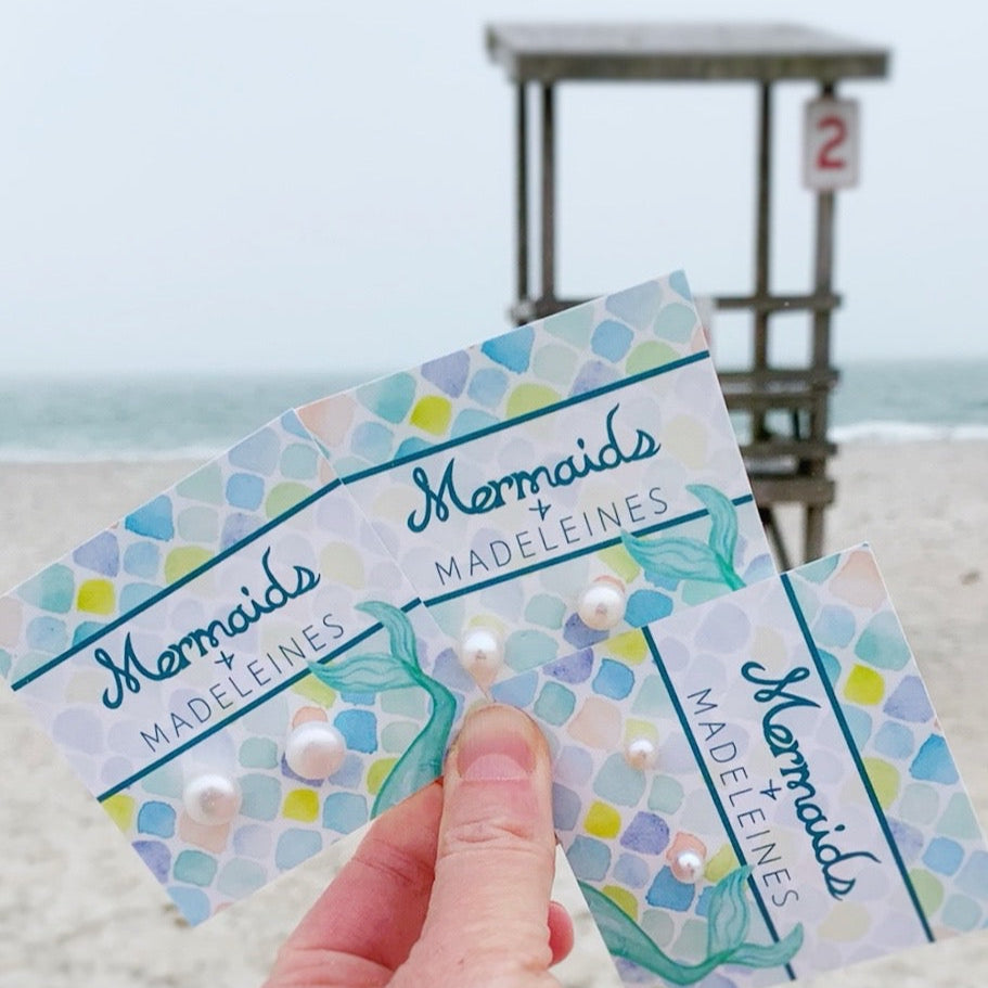 Pearlfect Day freshwater pearl post earrigns by mermaids and madeleines are available in 3 sizes, mini, daily and glam. Earrings are created with freshwater button pearls and sterling silver findings. This is a photographe of all 3 sizes carded held in a hand next to each other with a beach scene background
