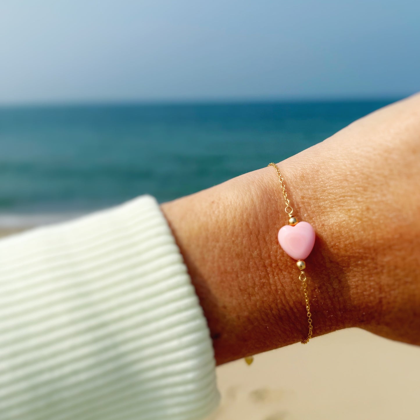 the pink posy adjustable bracelet by mermaids and madeleines is a chain style bracelet with a light pink, conch shell heart bead at the center. this bracelet is photographed up close on a wrist with a beach background