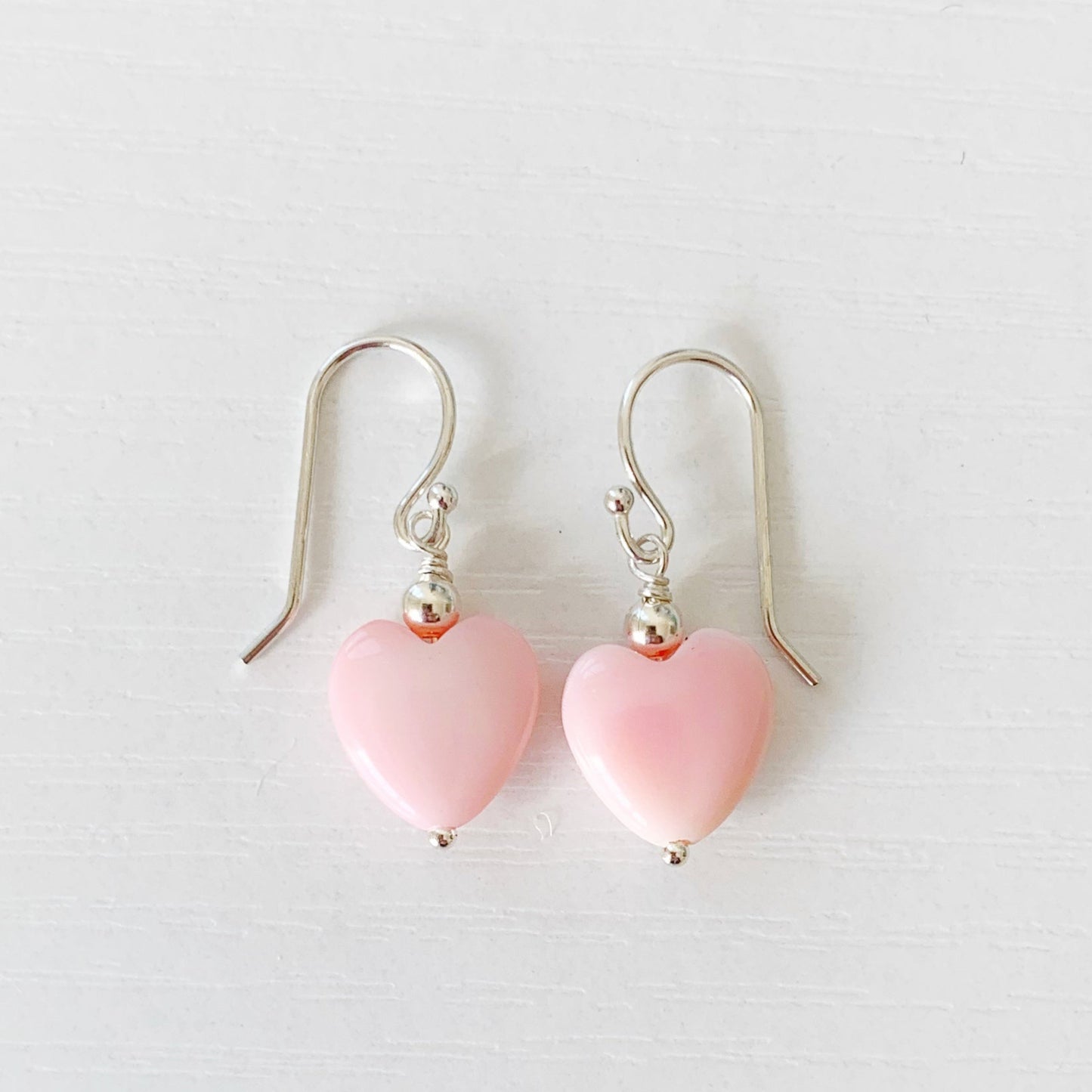 the pink posy earrings by mermaids and madeleines are queen conch shell carved hearts that are light pink. this pair has a sterling silver bead on top with sterling silver earring wires. The pair of earrings is photographed on a white surface.
