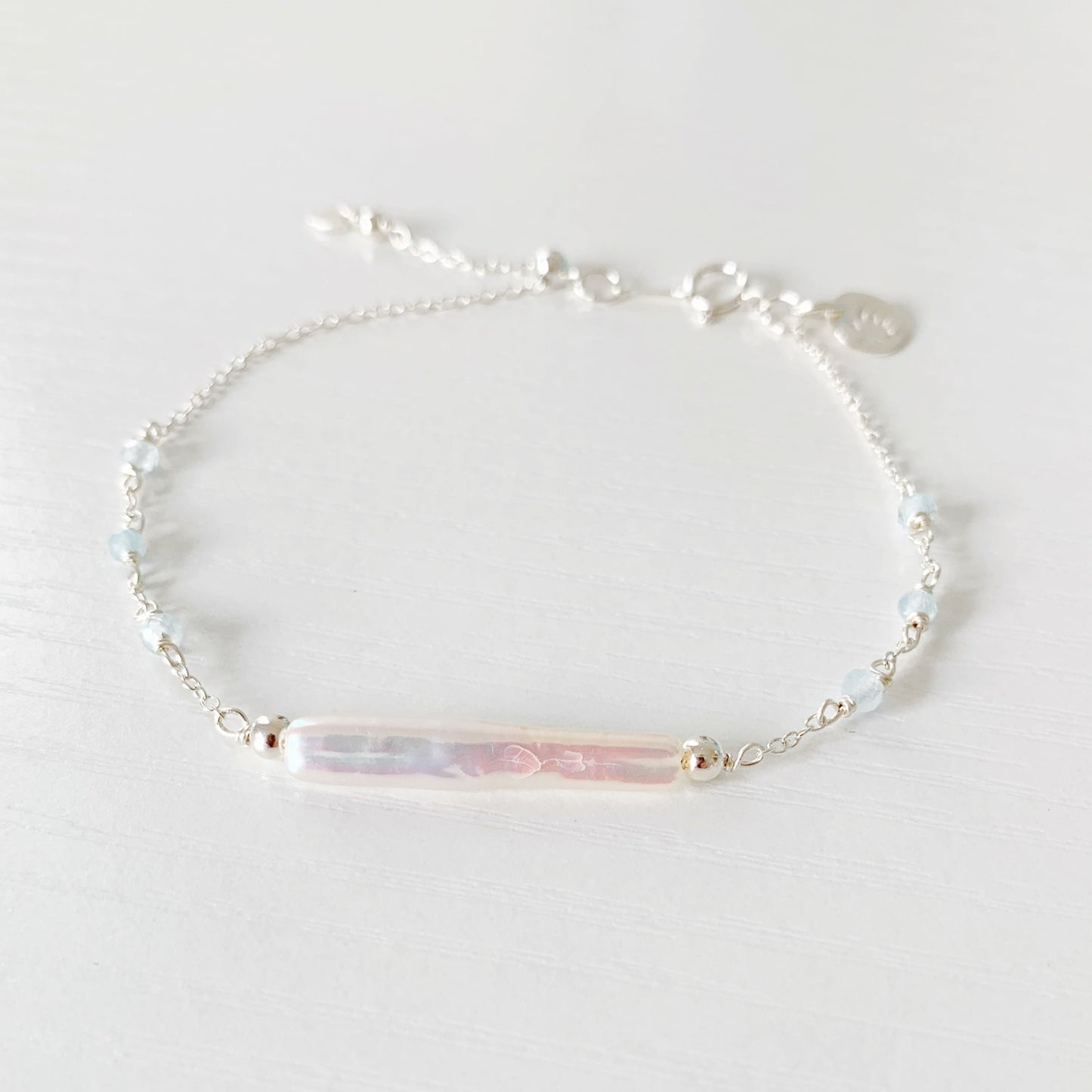the truro bracelet by mermaids and madeleines is a sterling silver chain bracelet with a freshwater stick pearl at the center and has small faceted aquamarine beads going down the sides with a slide bead near the clasp to make it adjustable. this bracelet is photographed flat on a white surface