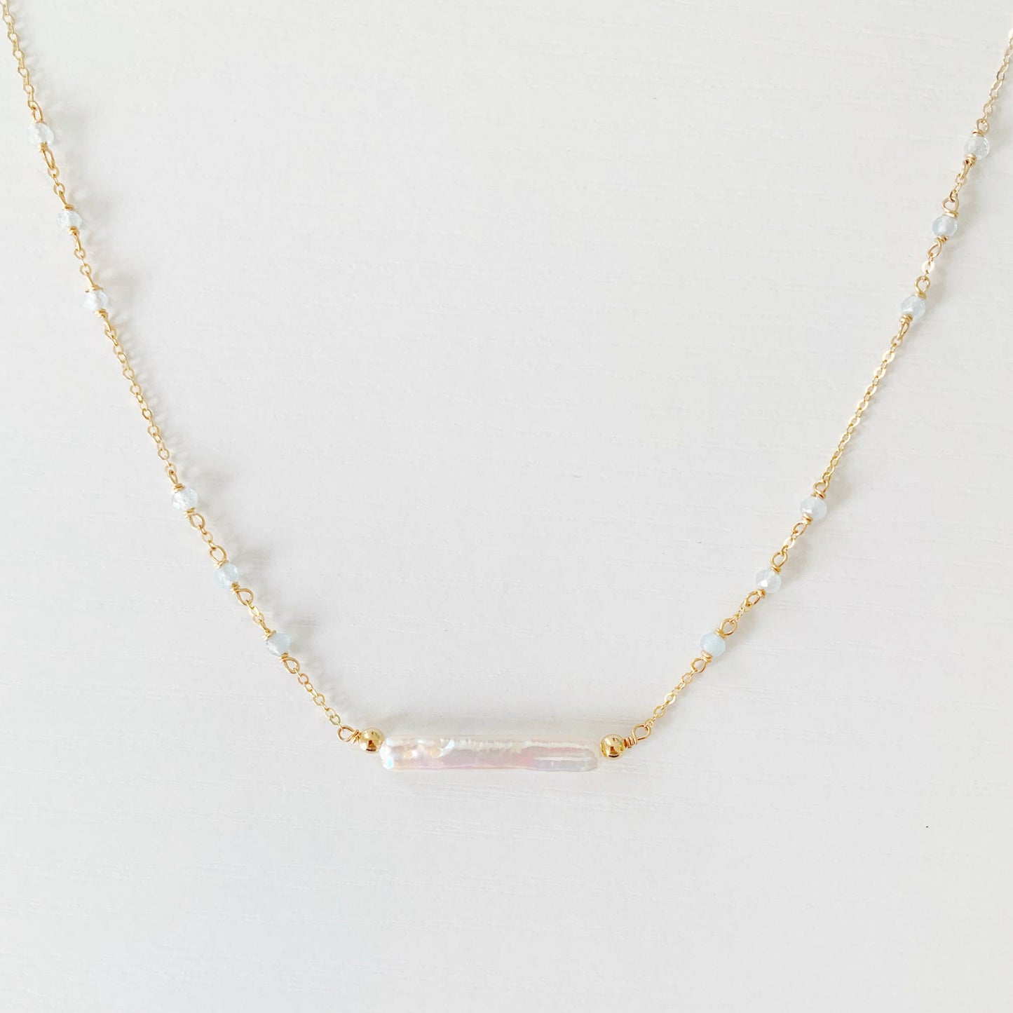 the truro necklace by mermaids and madeleines is a 14k gold filled chain necklace with a freshwater stick pearl at the center and aquamarine beads going up the sides. this necklace is photographed close up on a white surface
