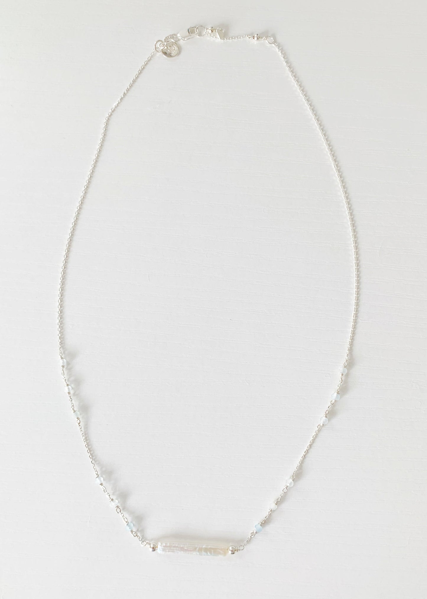 the truro necklace by mermaids and madeleines is a sterling silver chain necklace with a freshwater stick pearl in the center and aquamarine beads going up the sides. this necklace is photographed flat, with a full length view on a white surface