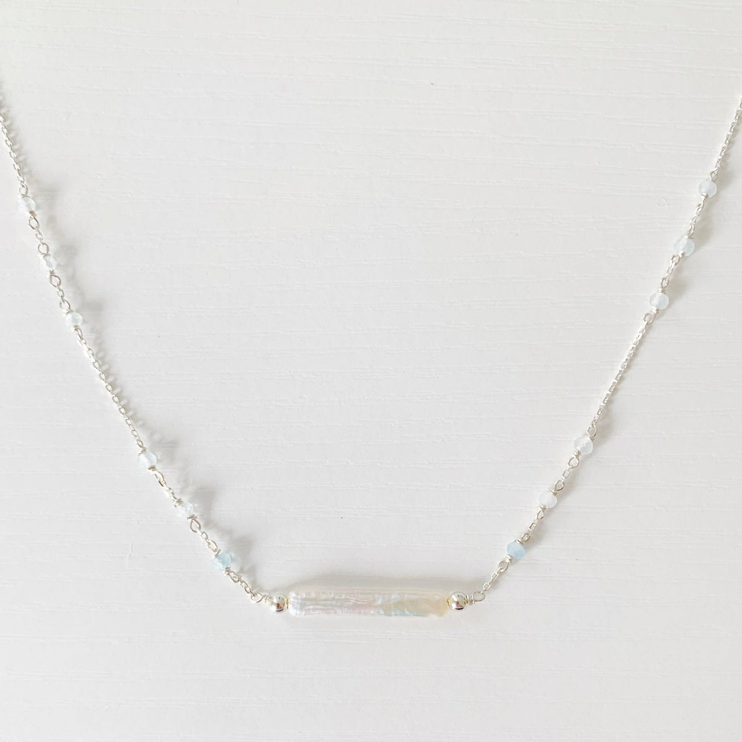 the truro necklace by mermaids and madeleines is a sterling silver chain necklace with a freshwater stick pearl at the center and aquamarine beads going up the sides of the chain. this necklace is photographed close up on a white surface