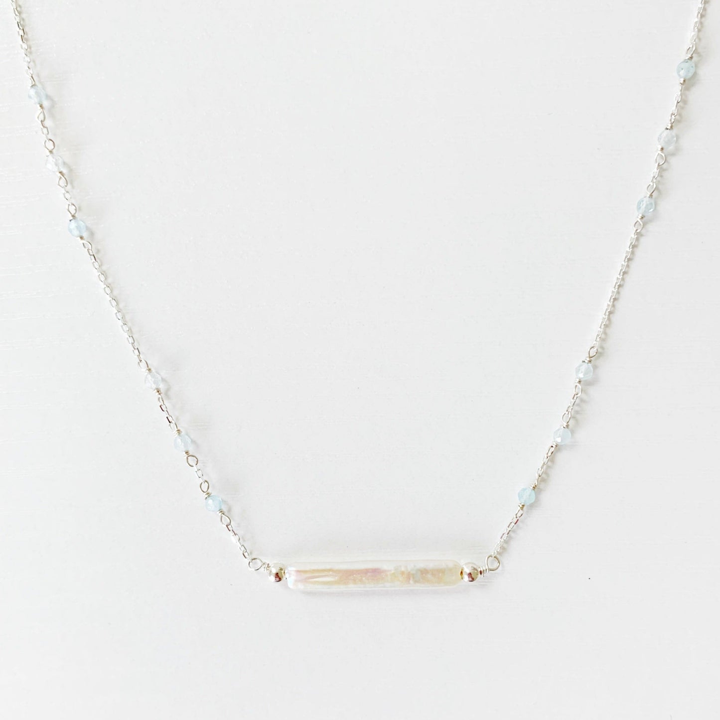 The Truro Necklace by Mermaids and Madeleines features a freshwater stick pearl at the center like a horizontal bar necklace with tiny microfaceted aquamarine beads going up the chain on either side. the chain and findings are sterling silver. this necklace is photographed on a white surface