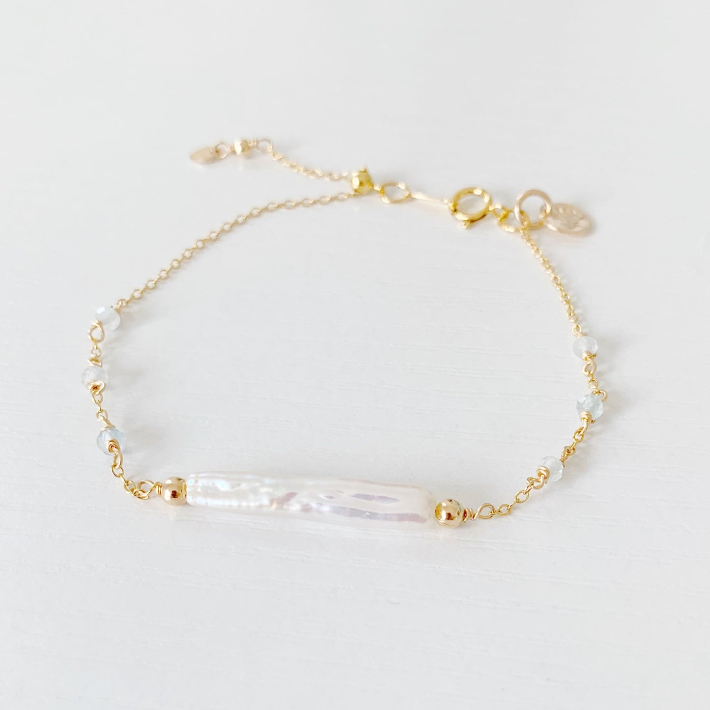 the truro bracelet by mermaids and madeleines is a 14k gold filled chain bracelet with a freshwater stick pearl at the center and aquamarine faceted beads going down the sides. there is a slide bead near the clasp to make the bracelet adjustable. this bracelet is photographed flat on a white surface