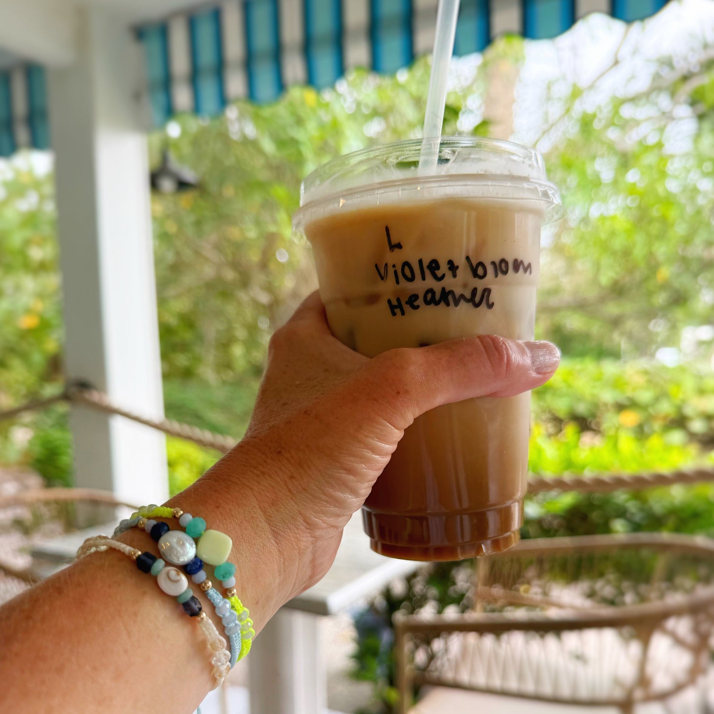 Hand holding a iced coffee drink with a straw, wearing bracelets, on a porch.