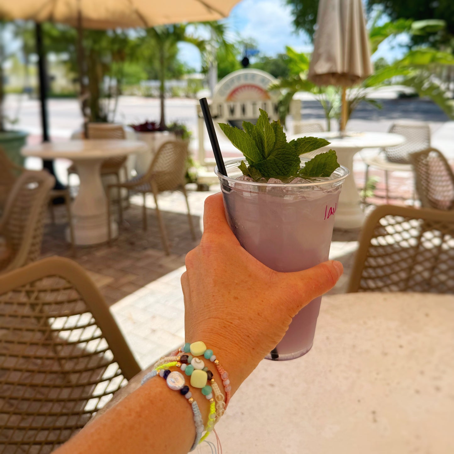 Hand holding a pink drink with a straw and mint leaf in an outdoor setting with tables and chairs.