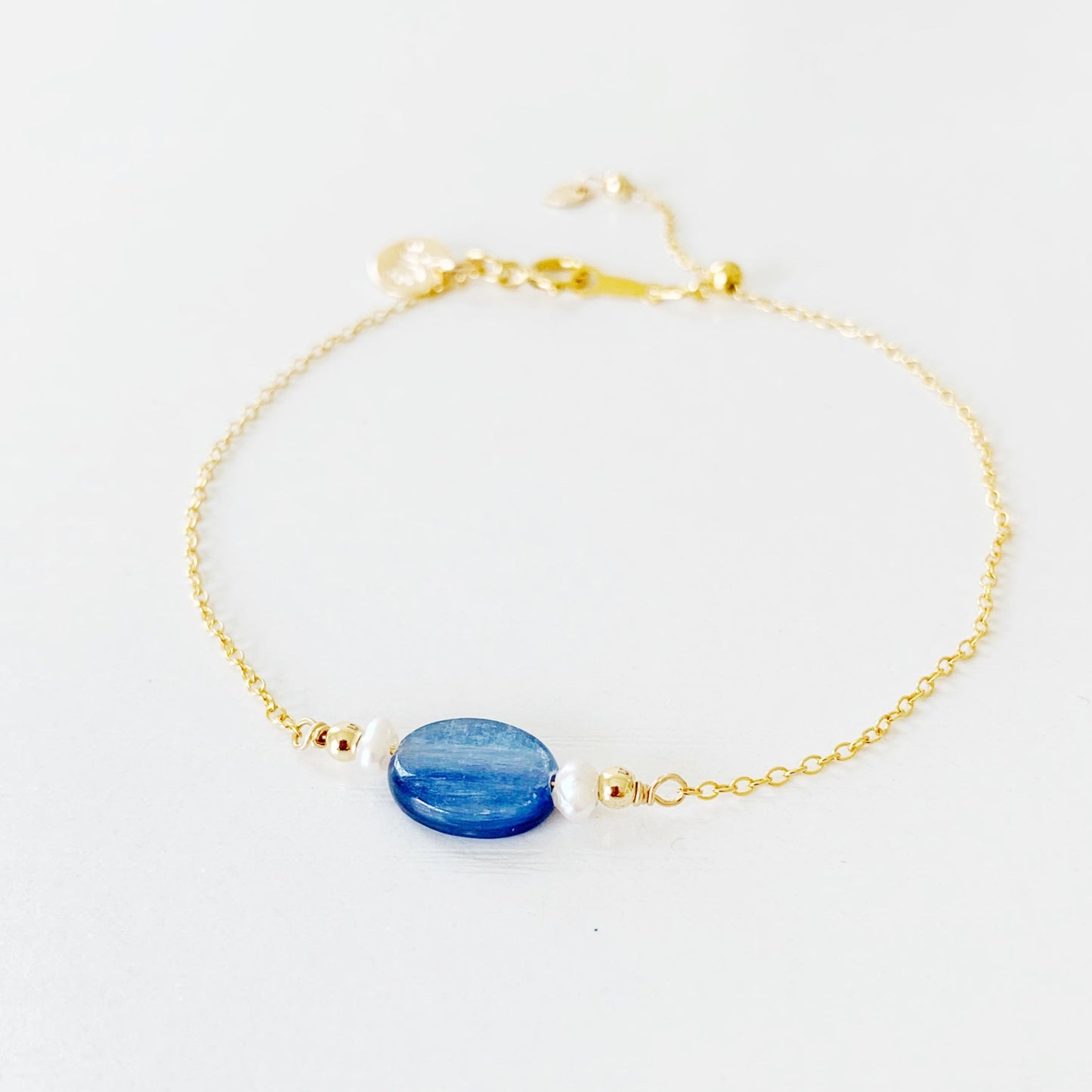 the brewster bracelet by mermaids and madeleines is an adjustable 14k gold filled chain bracelet with a slide bead at the clasp. there's a blue kyanite coin bead at the center complimented by small freshwater pearls on each side. this is picture of the brewster bracelet photographed on a white surface