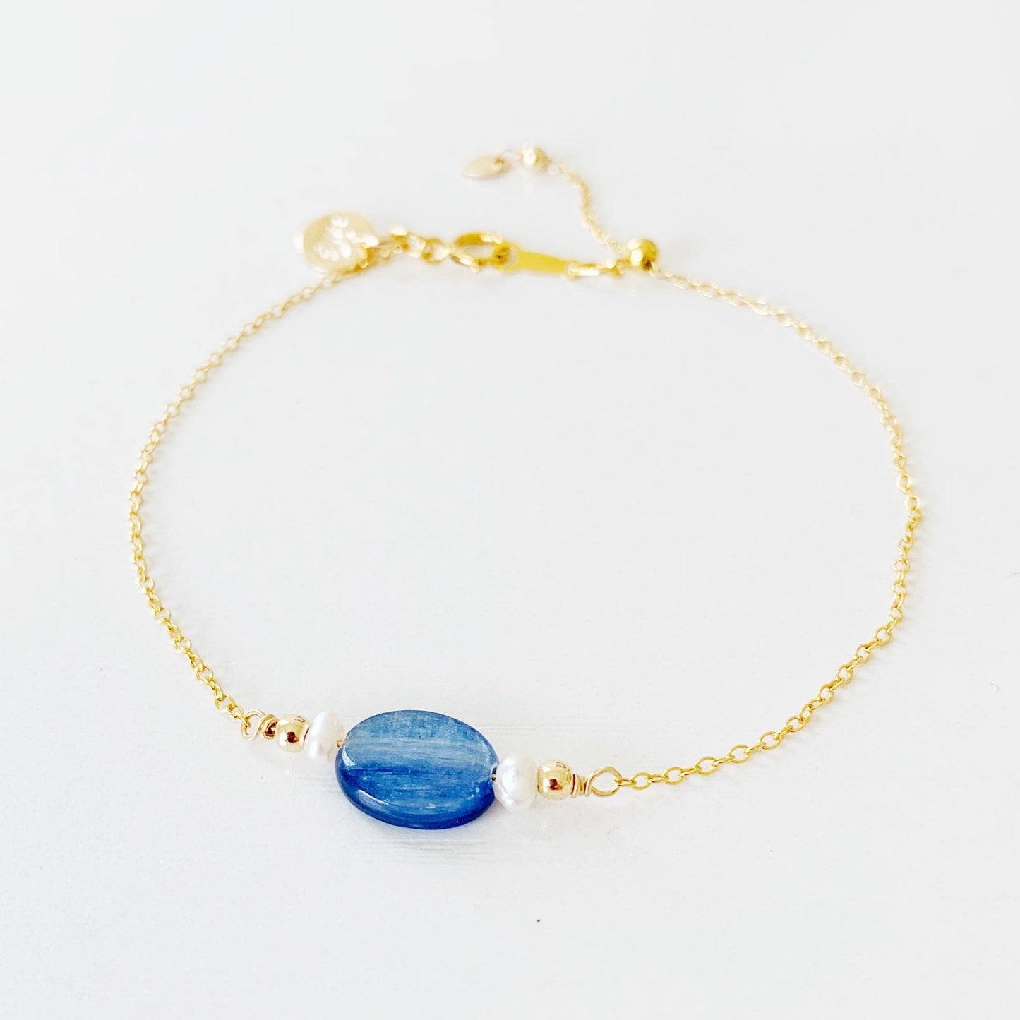 the brewster bracelet by mermaids and madeleines is an adjustable 14k gold filled chain bracelet with a slide bead at the clasp. there's a blue kyanite coin bead at the center complimented by small freshwater pearls on each side. this is a photo of the brewster bracelet photographed flat on a white surface