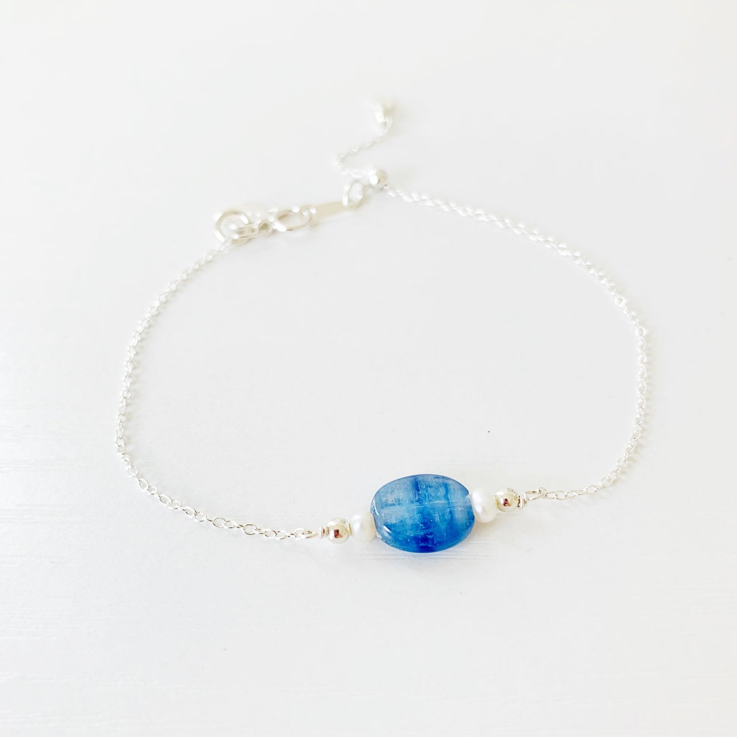 the brewster bracelet by mermaids and madeleines is an adjustable sterling silver chain bracelet with a slide bead at the clasp. there's a blue kyanite coin bead at the center complimented by small freshwater pearls on each side. this bracelet is photographed from a front view on a white surface