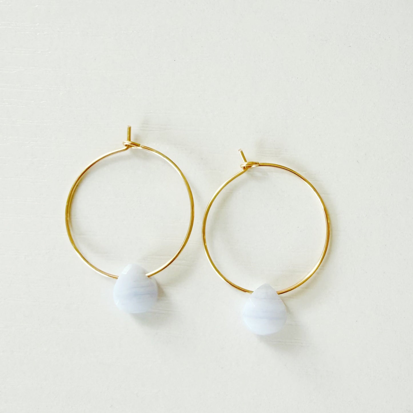 A pair of gold hoop earrings with a small pastel purple bead dangling from the bottom, displayed on a clean white background.