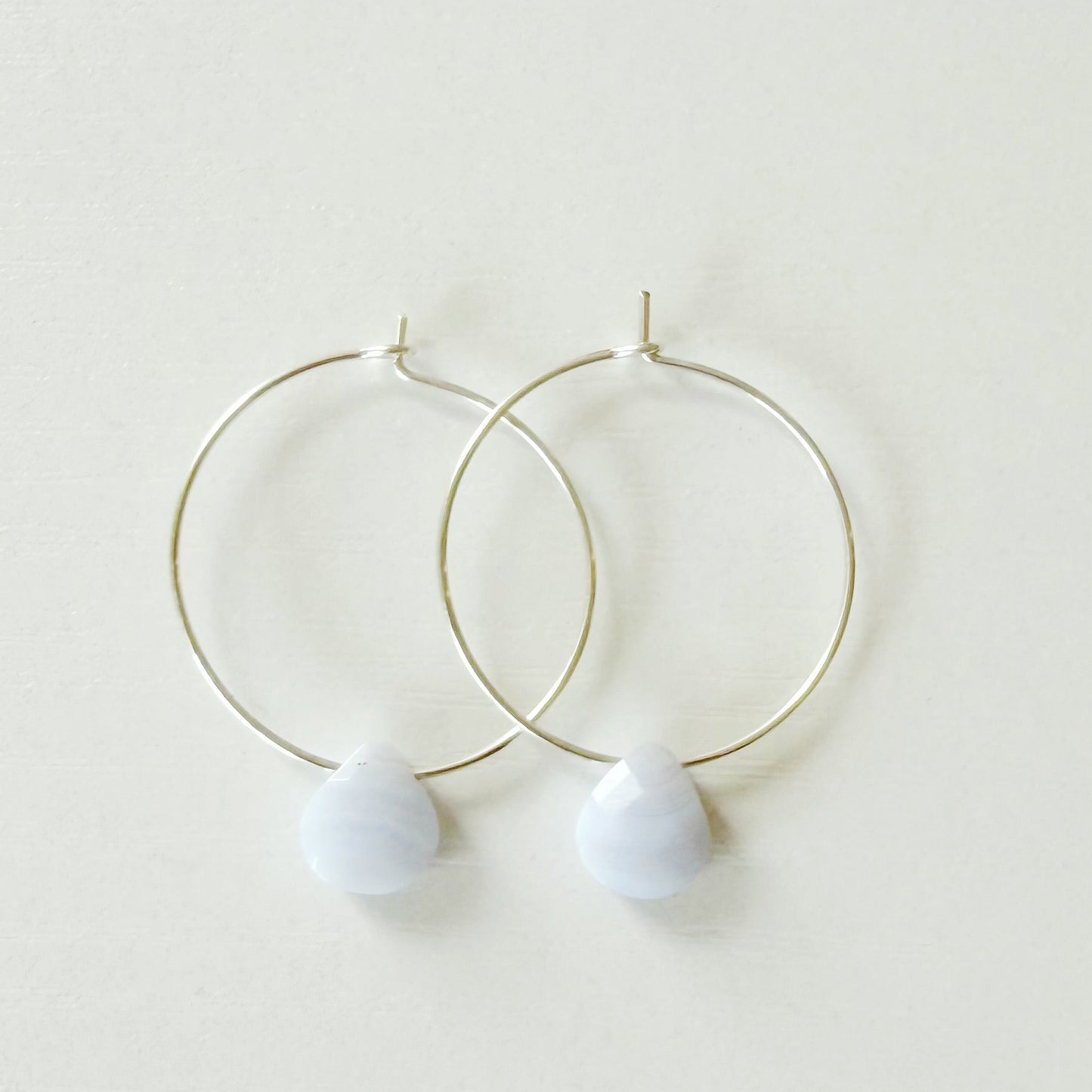 A pair of delicate silver hoop earrings with small, teardrop-shaped pastel purple beads hanging from the bottom, displayed on a clean, off-white background.