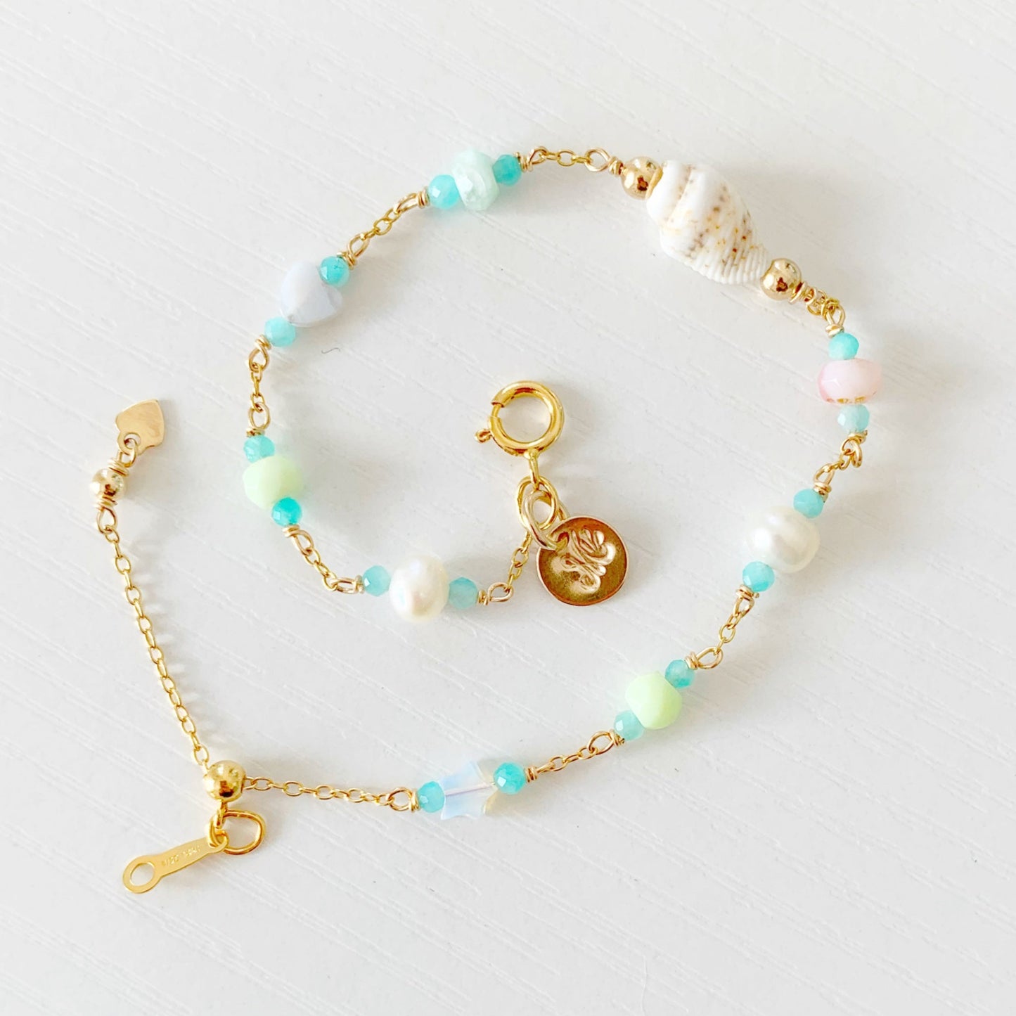 the island hopper adjustable bracelet by mermaids and madeleines is a station style bracelet with a shell at the center and semiprecious beads going up the sides of the 14k gold filled chain. this bracelet is laid out in a spiral shape and photographed from the top down on a white surface