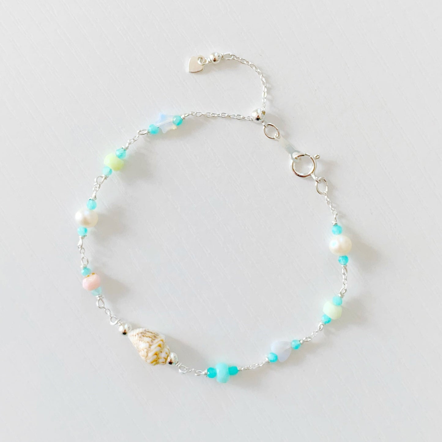 the island hopper adjustable bracelet by mermaids and madeleines is a station style bracelet with a shell at the center and semiprecious beads going up the sterling silver chain. the bracelet has a slide bead near the clasp making it adjustable. this bracelet is photographed from a top down view on a white surface