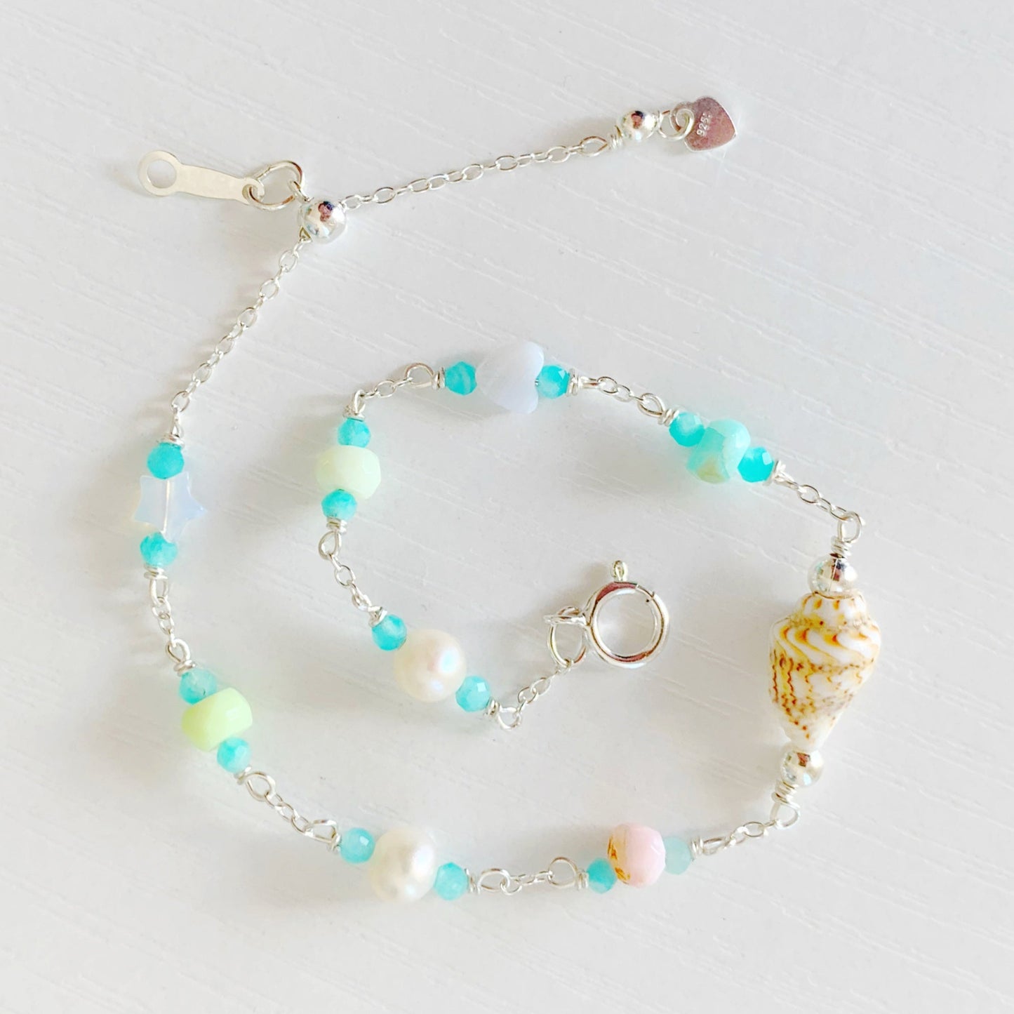 the island hopper bracelet by mermaids and madeleines is a station style bracelet with a shell at the center and semiprecious beads going up the sterling chain. this bracelet is photographed from the top down with the bracelet in a spiral shap on a white surface