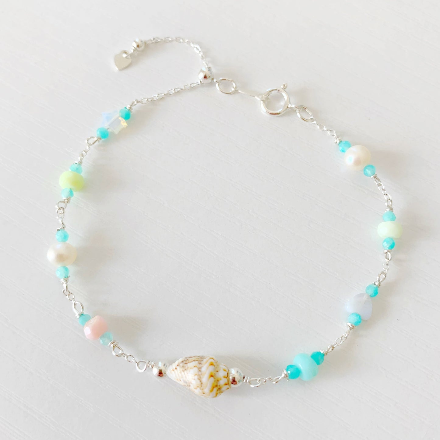the island hopper bracelet by mermaids and madeleines is an adjustable station style bracelet with a shell at the center with semiprecious beads going up the sterling chain. this bracelet is photographed from the top down on a white surface