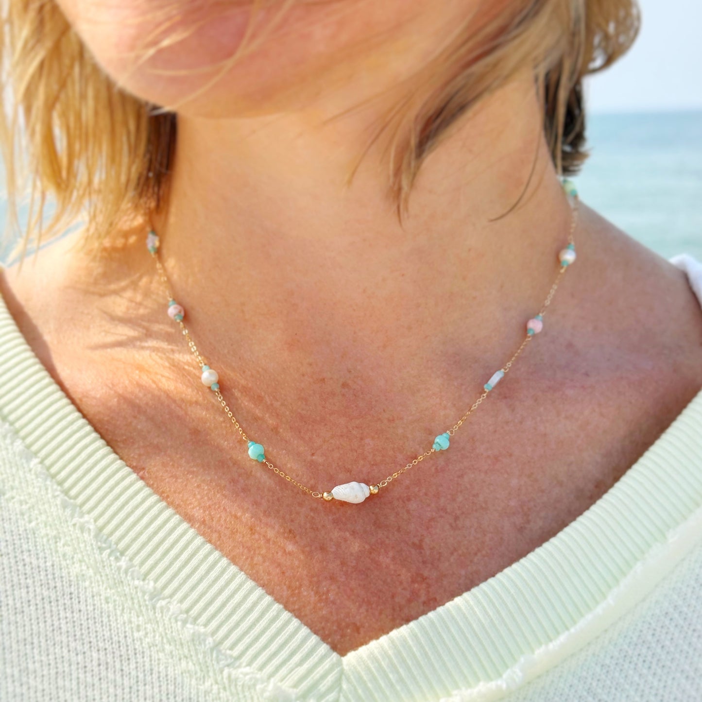 the island hopper necklace by mermaids and madeleines is a station style necklace with a shell at the center and semiprecious beads going up the 14k gold filled chain. this necklace is photographed close up on a person with a beach background