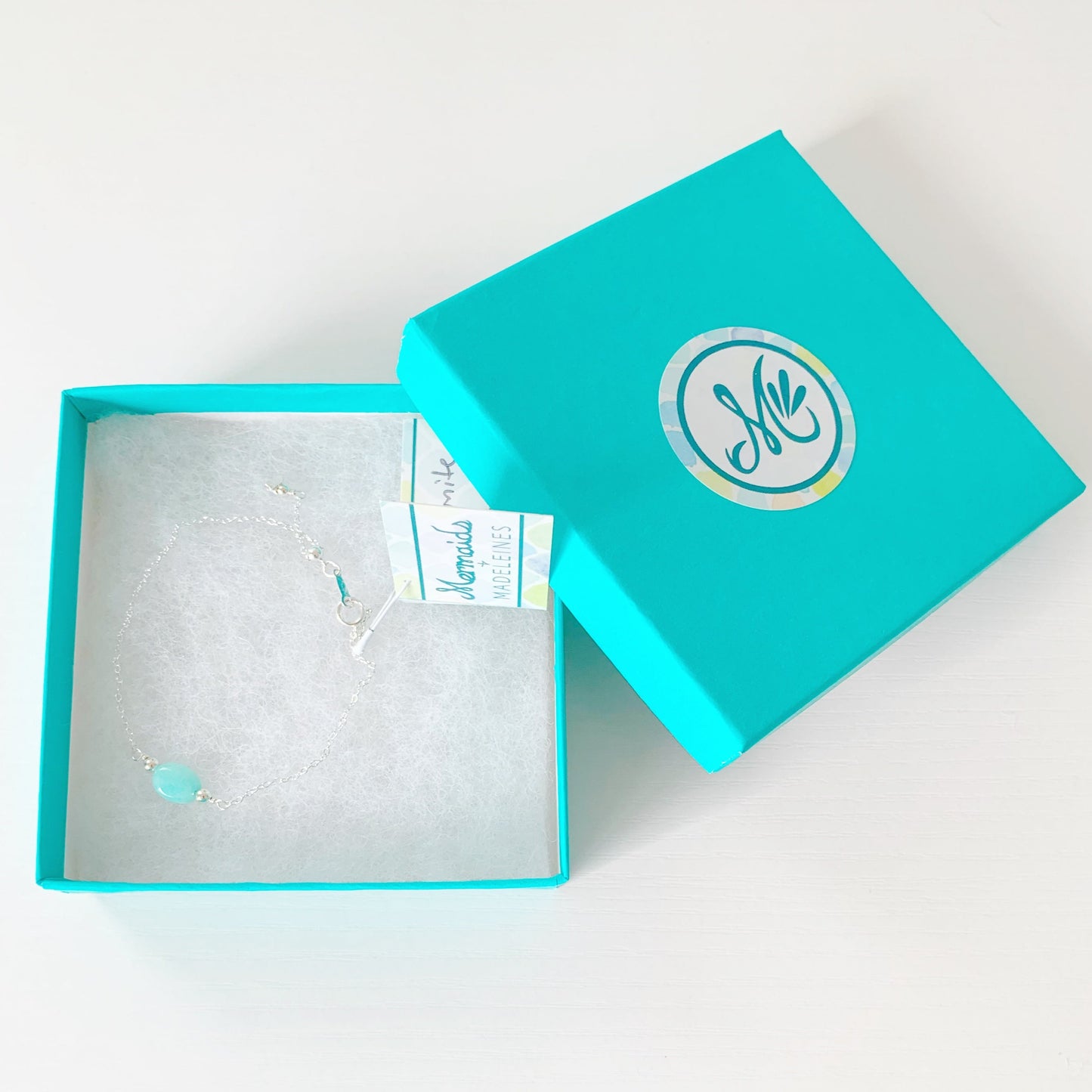 the laguna adjustable bracelet by mermaids and madeleines is created with sterling silver chain with an aqua oval amazonite bead at the center. There's a slide bead at the clasp to make the chain bracelet adjustable. this bracelet is photographed in a teal gift box on a white surface