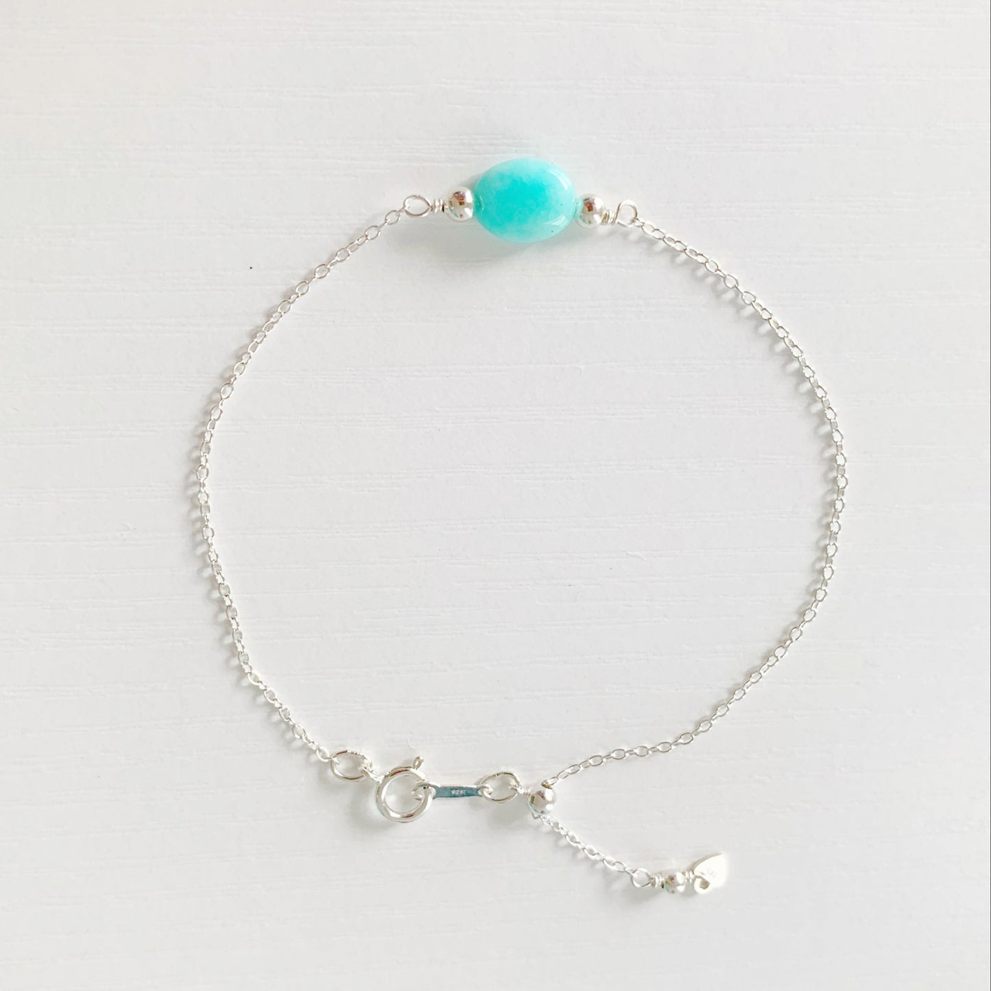 the laguna bracelet by mermaids and madeleines is a sterling silver chain based bracelet with an aqua amazonite oval bead a the center and with sterling slide bead near the clasp making it adjustable. this bracelet is photographed from the top down on a white surface