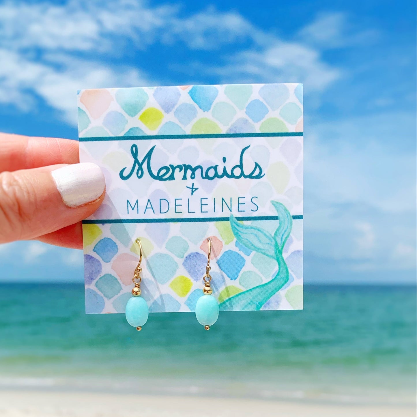 the laguna earrings by mermaids and madeleines are amazonite oval drops suspended from 14k gold filled findings and ear wires. this pair is carded and held up in front of a beach scene