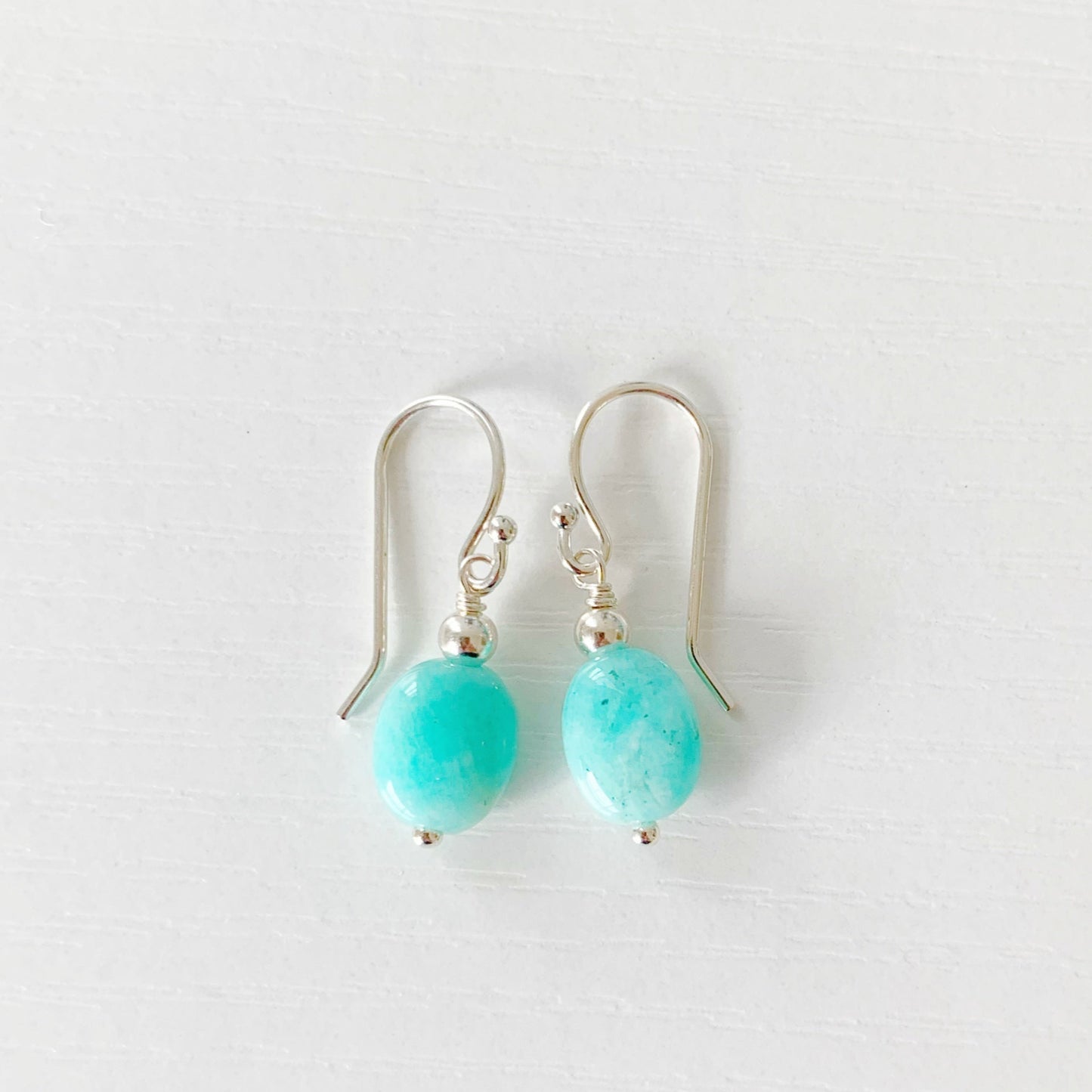 the laguna earrings by mermaids and madeleines are simple drop style earrings with bright aqua amazonite oval beads dropped from sterling silver findings and ear wires. this pair of earrings are photographed on a white surface
