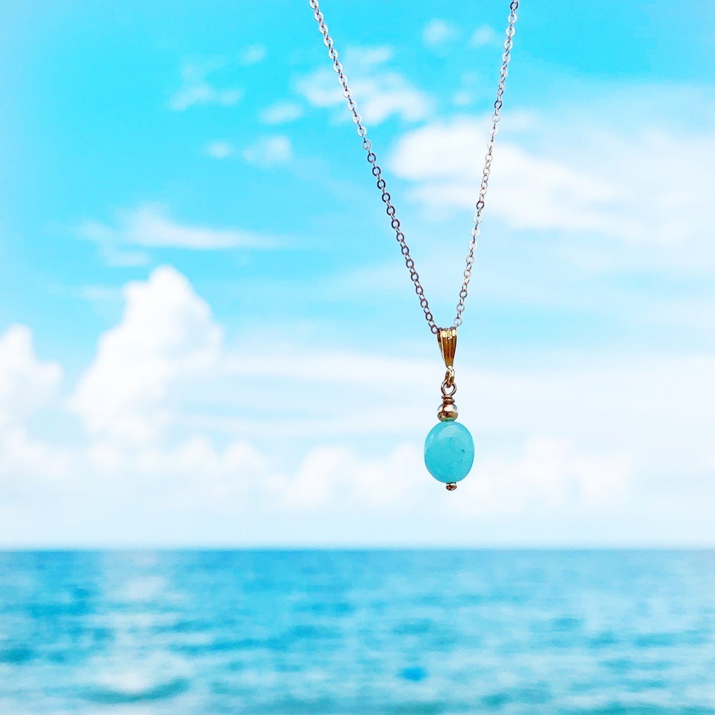 Laguna necklace by mermaids and madeleines is created with an aqua color amazonite oval stone and hangs from a 14k gold filled chain. this necklace is pictured suspenced over a blue sky and beachy background